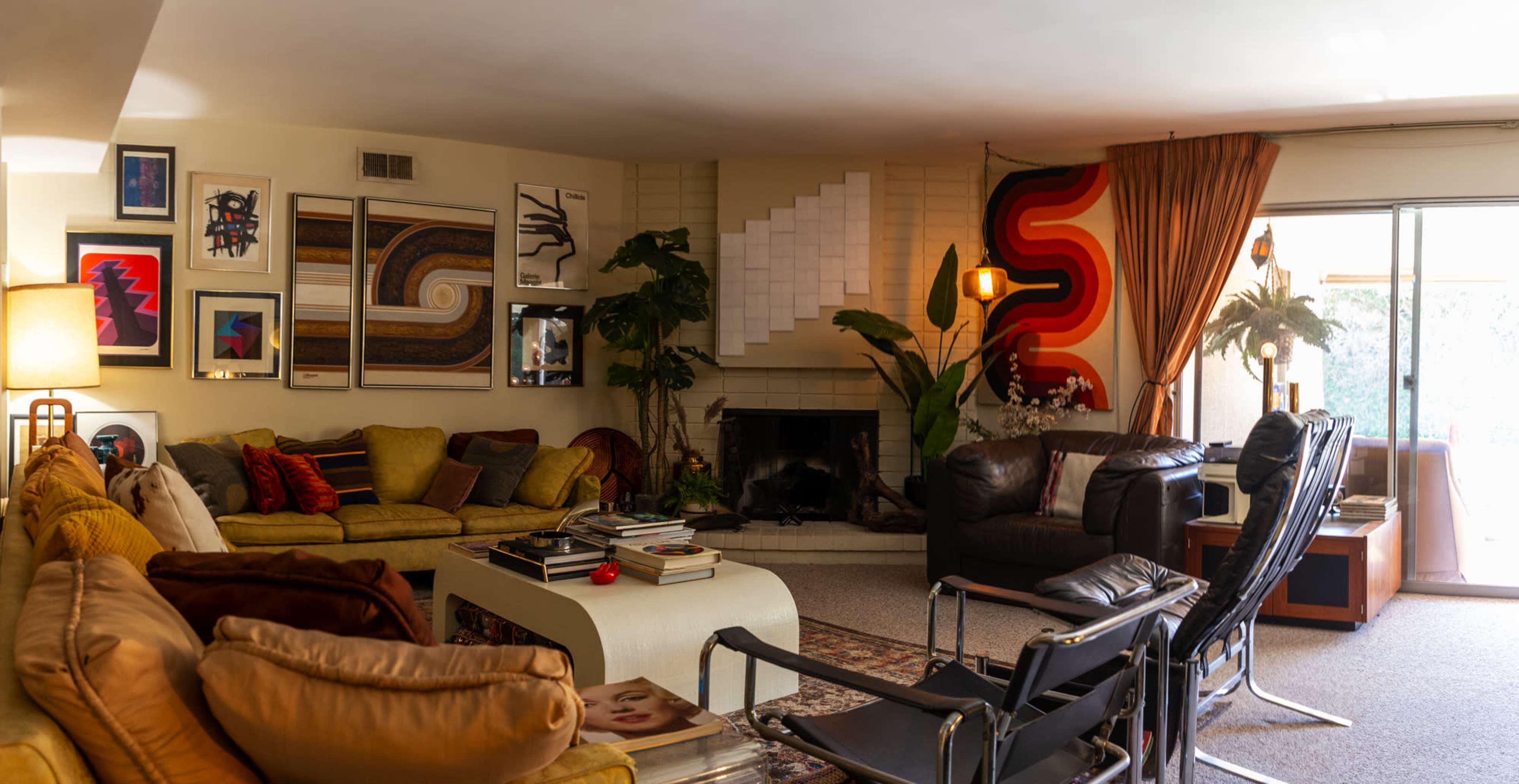 A spacious living room features a large sectional sofa, a fireplace with a mantel, various pieces of wall art, and a mix of plants and furniture including a reclining chair and a coffee table.