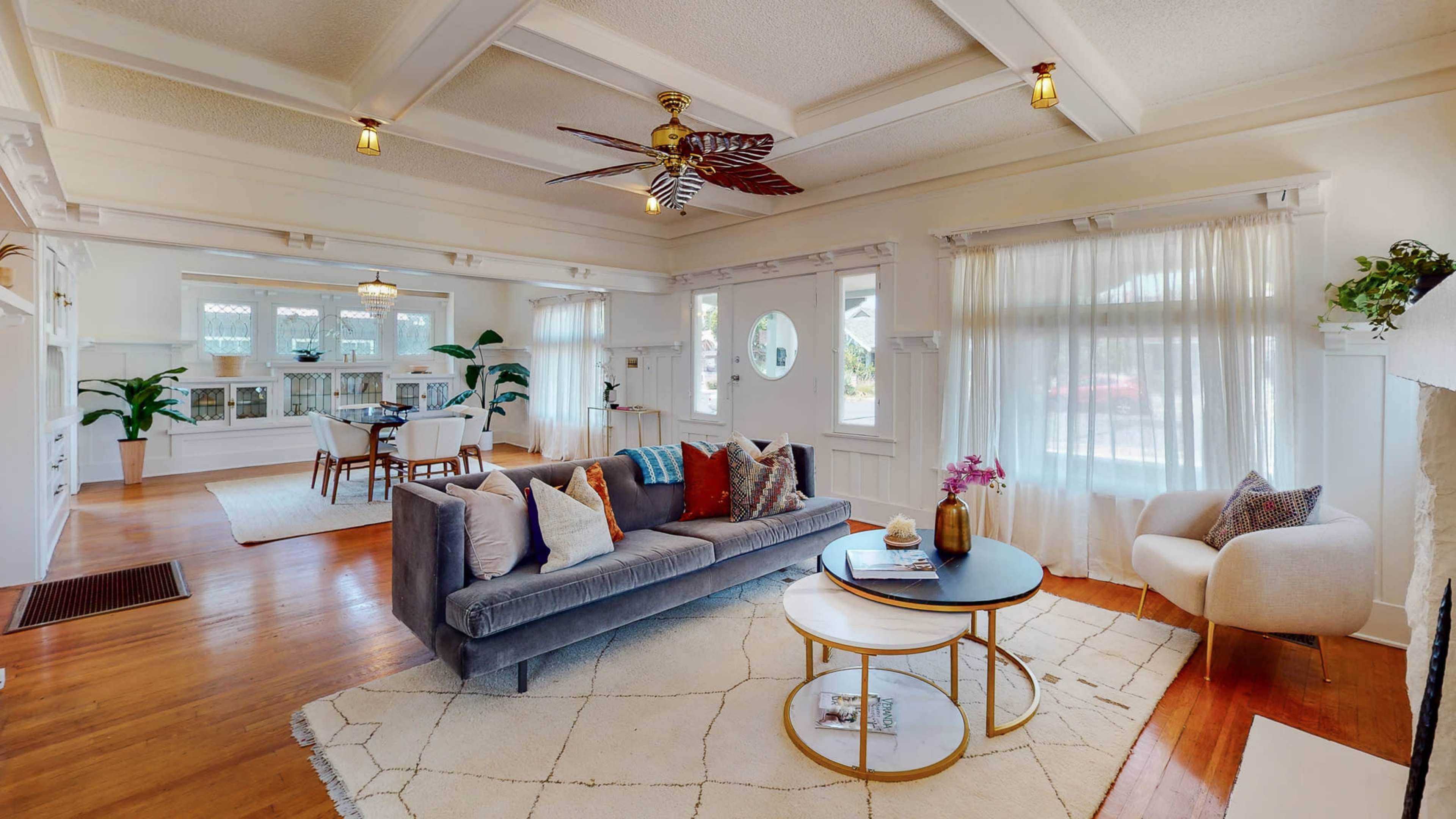 The image shows a bright living room with a gray sofa, a round coffee table, and a dining area featuring a table and chairs, all set within an elegantly designed space with white walls and wooden accents.