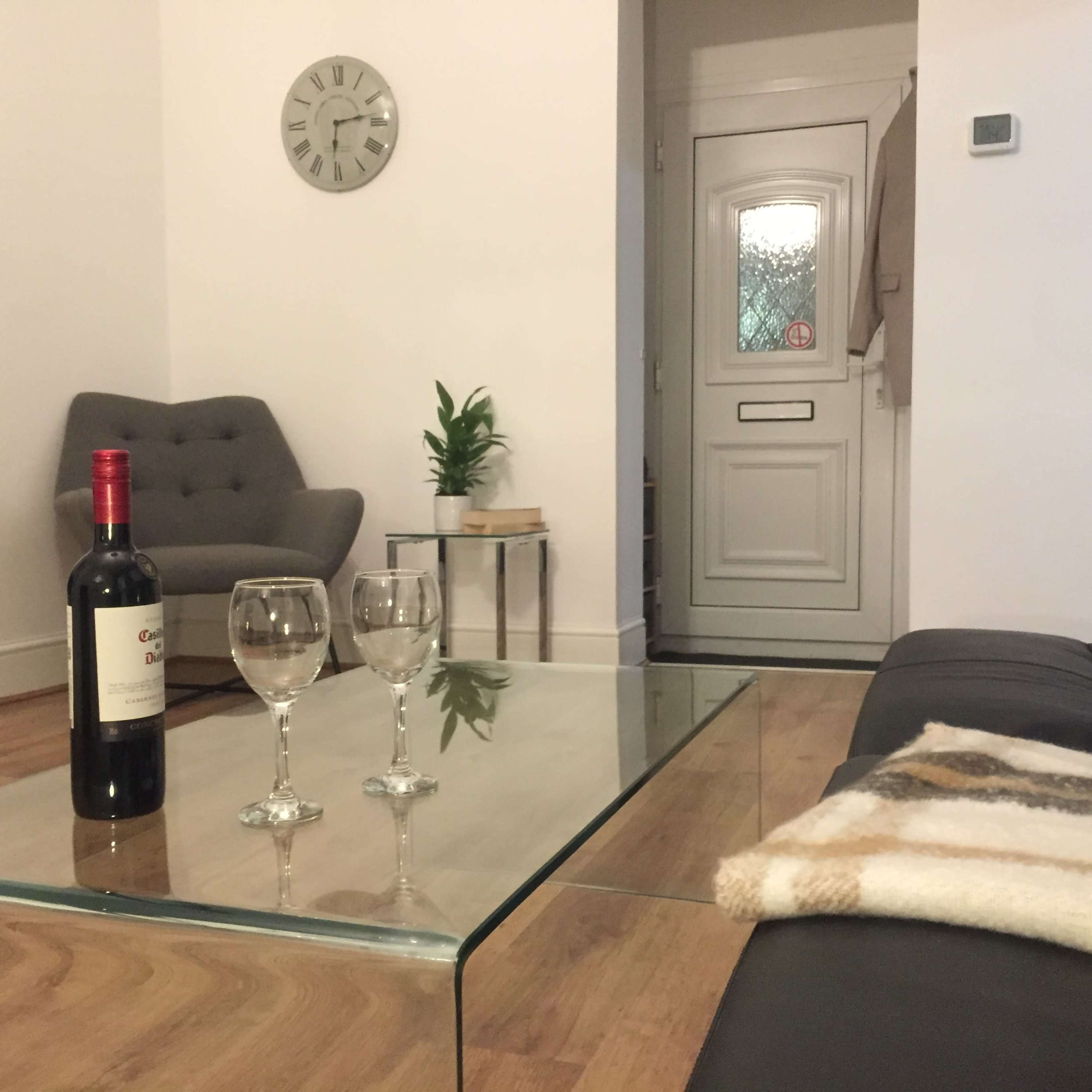 A modern living room features a glass coffee table with two wine glasses and a bottle of wine, alongside a sofa and a view of the entrance door.