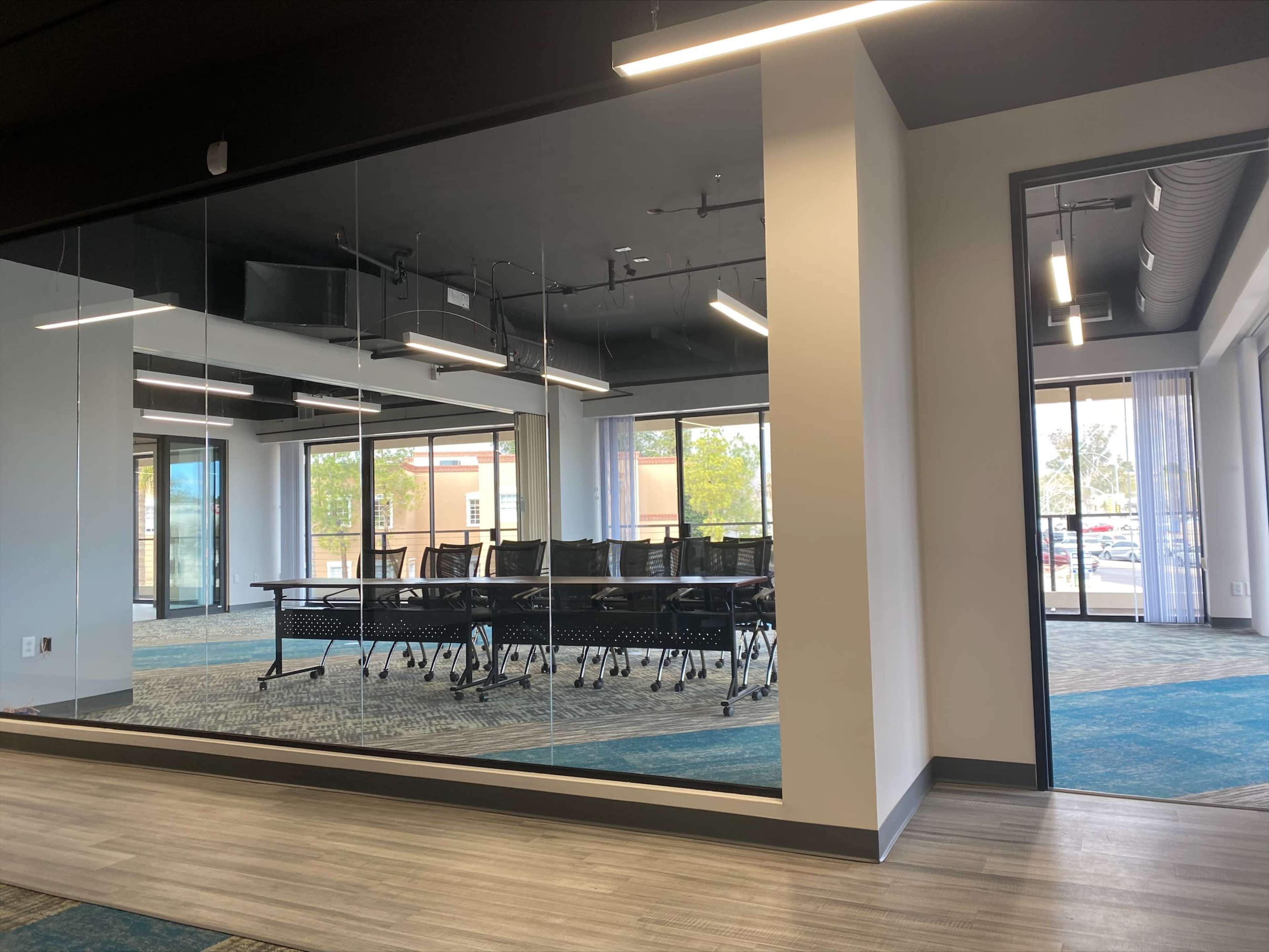 A modern conference room with a glass wall, featuring a long table and multiple chairs, visible from an adjacent hallway.