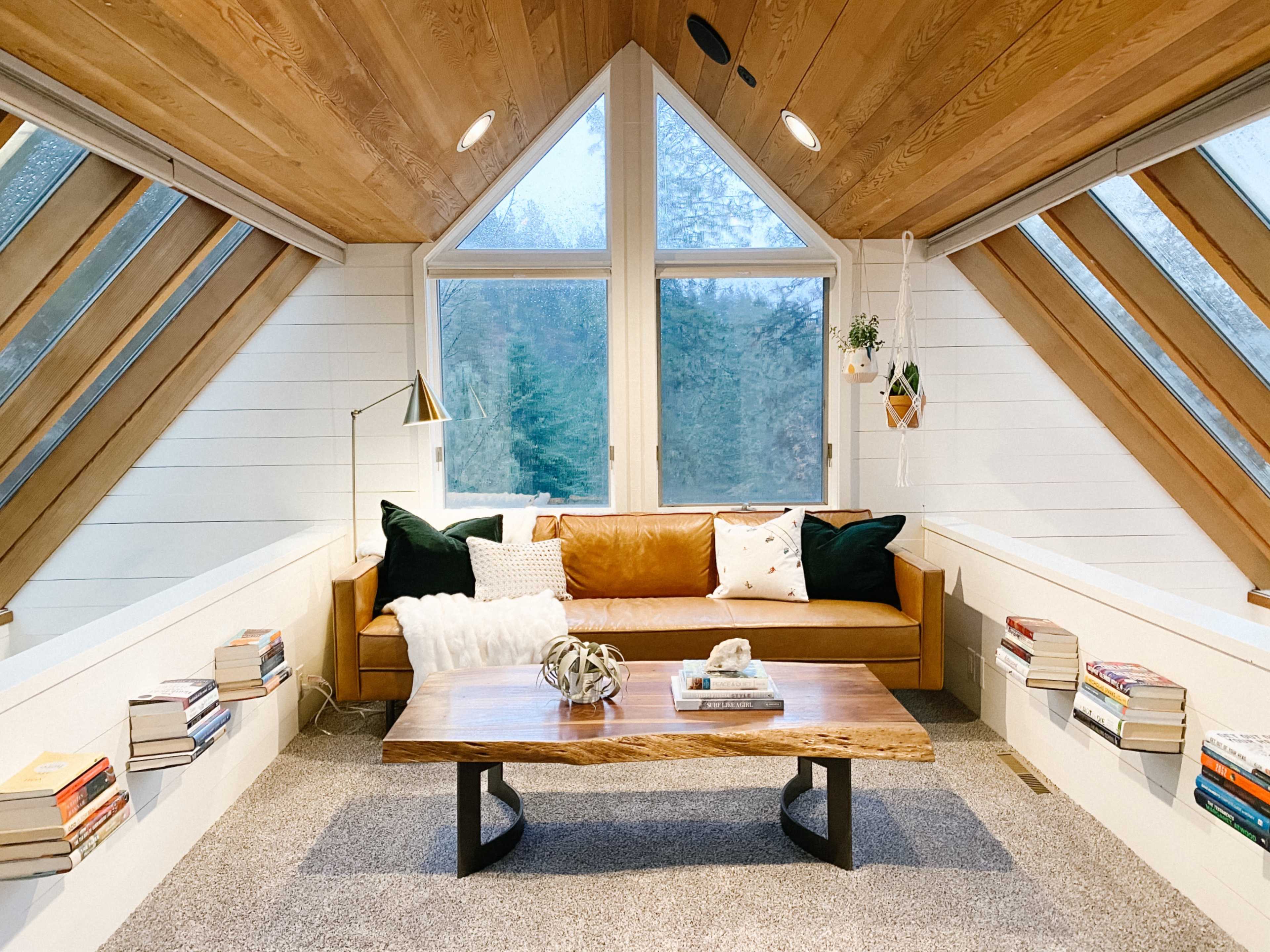 A cozy loft space features a brown leather sofa, a rustic wooden coffee table, and large windows with angled roofs, surrounded by bookshelves built into the walls.