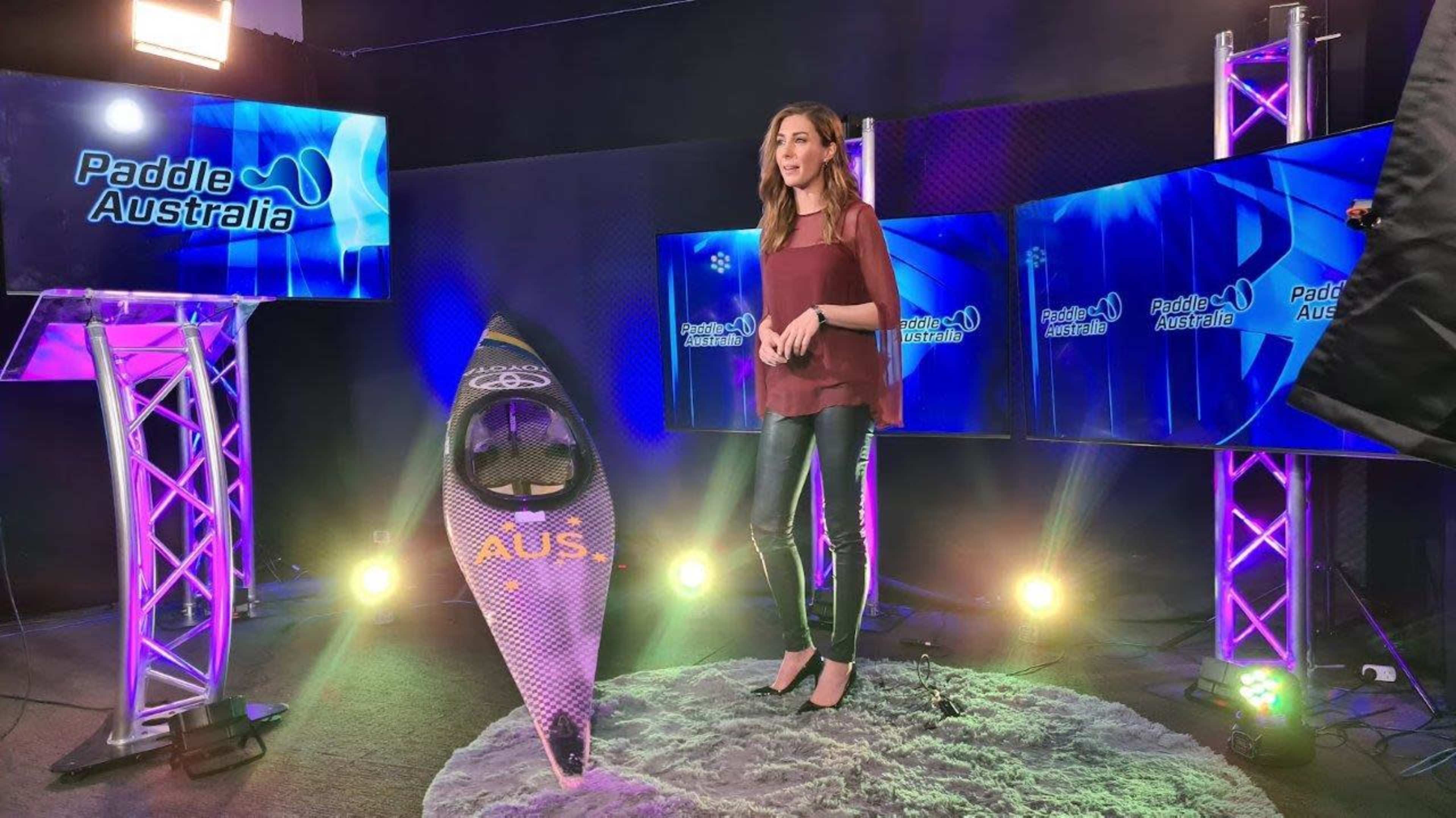 A presenter stands on a stage with multiple screens displaying "Paddle Australia" and a kayak nearby.