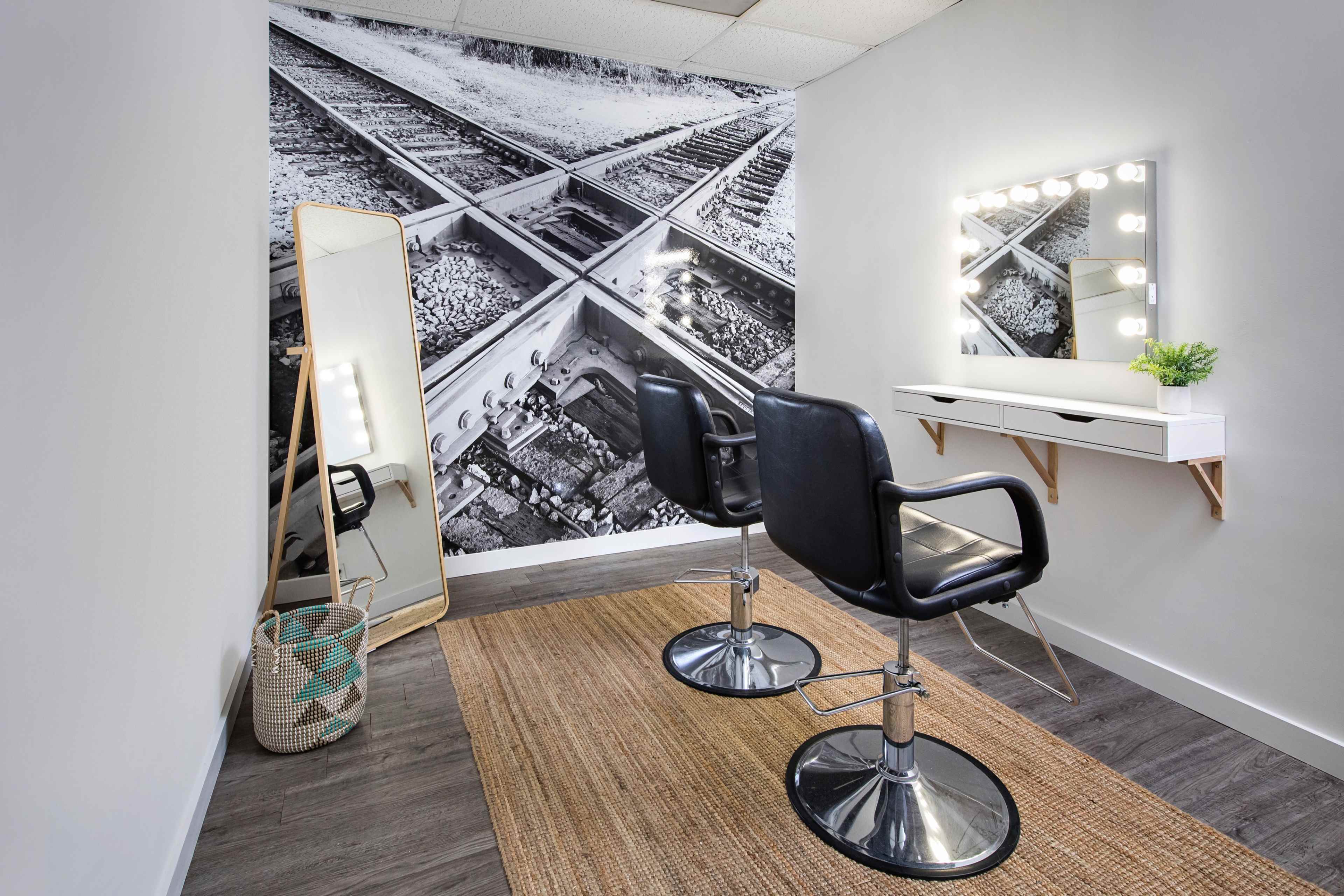 A minimalist salon interior featuring two black chairs, a wall-mounted mirror with lights, and a large black-and-white mural of railway tracks.