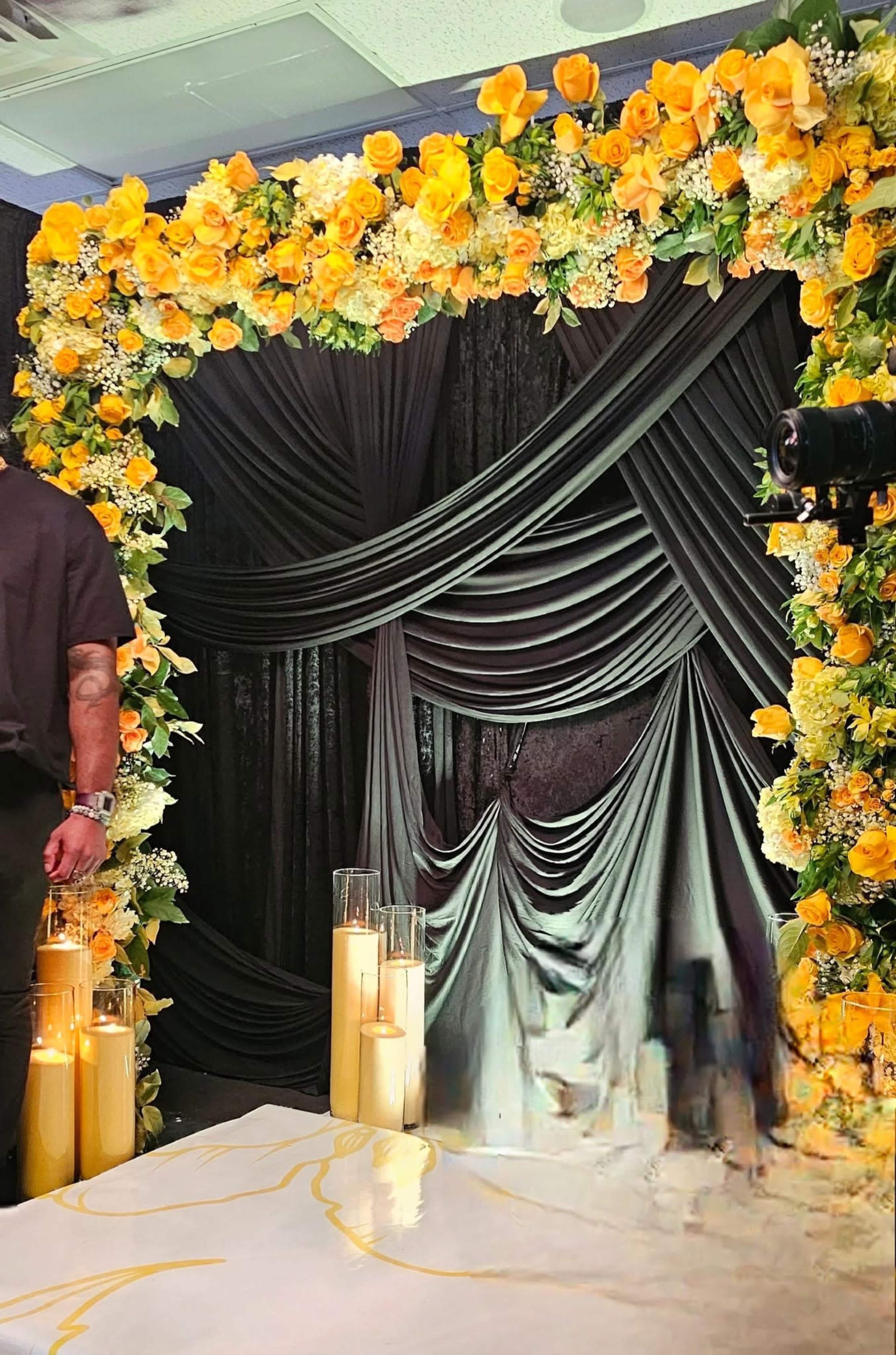 A photo booth backdrop is decorated with a floral arrangement of yellow roses and greenery, framed by flowing black fabric and candles placed on the floor.