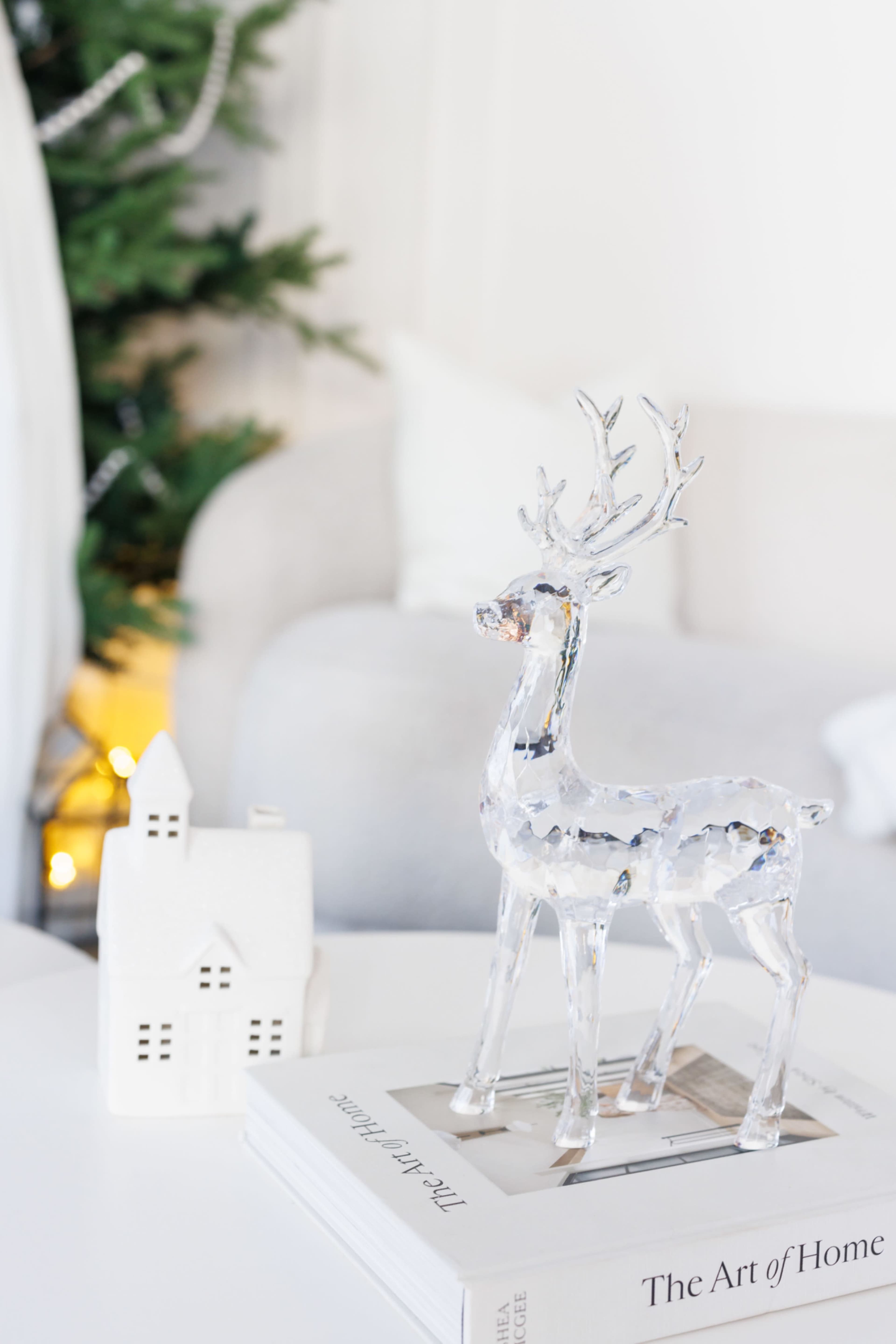 A clear crystal reindeer figurine stands on a stacked coffee table book beside a small white house decoration, with a softly lit Christmas tree in the background.