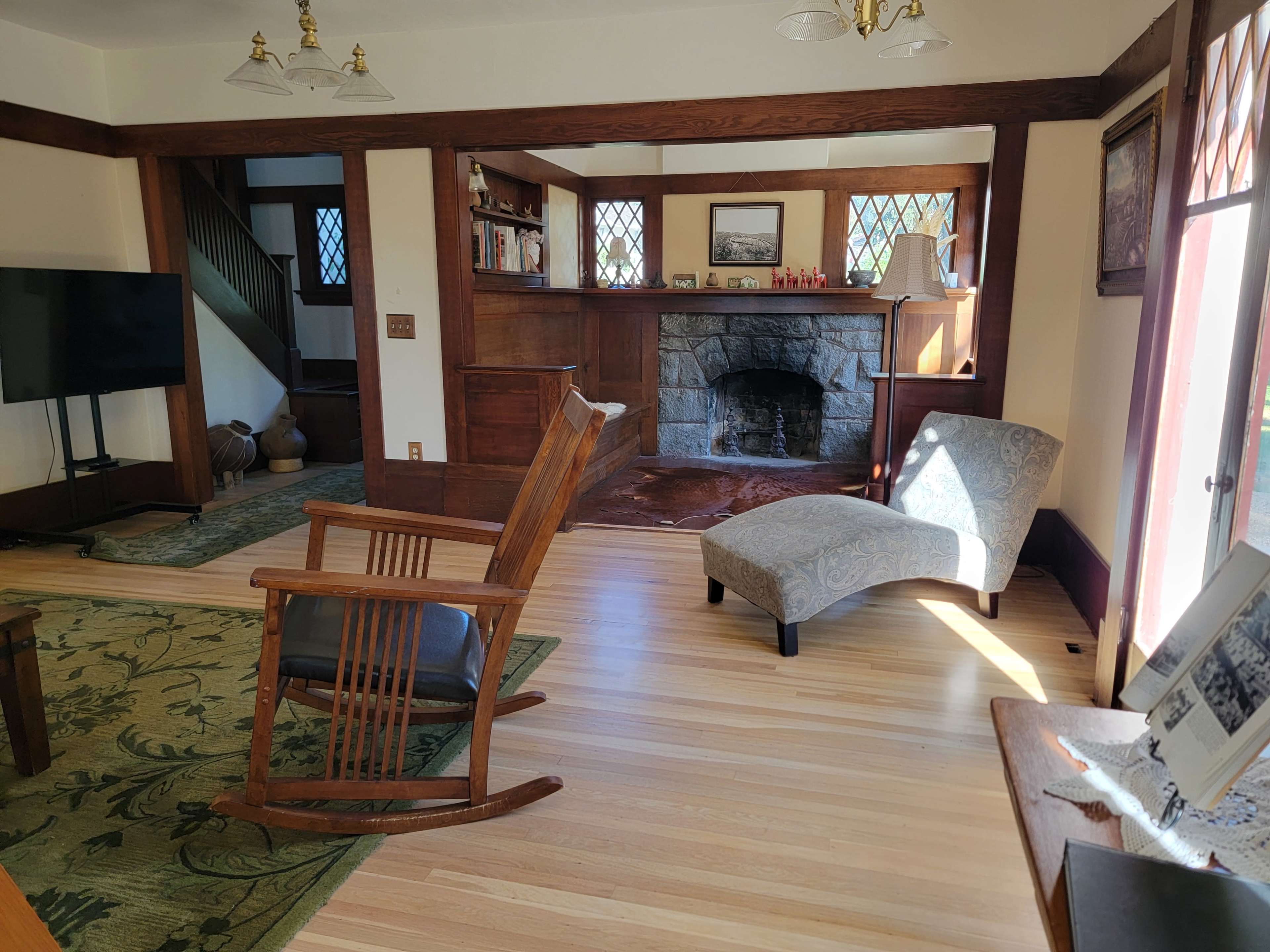 The room features a rocking chair, a patterned chaise lounge, a stone fireplace, wooden accents, and large windows allowing natural light.