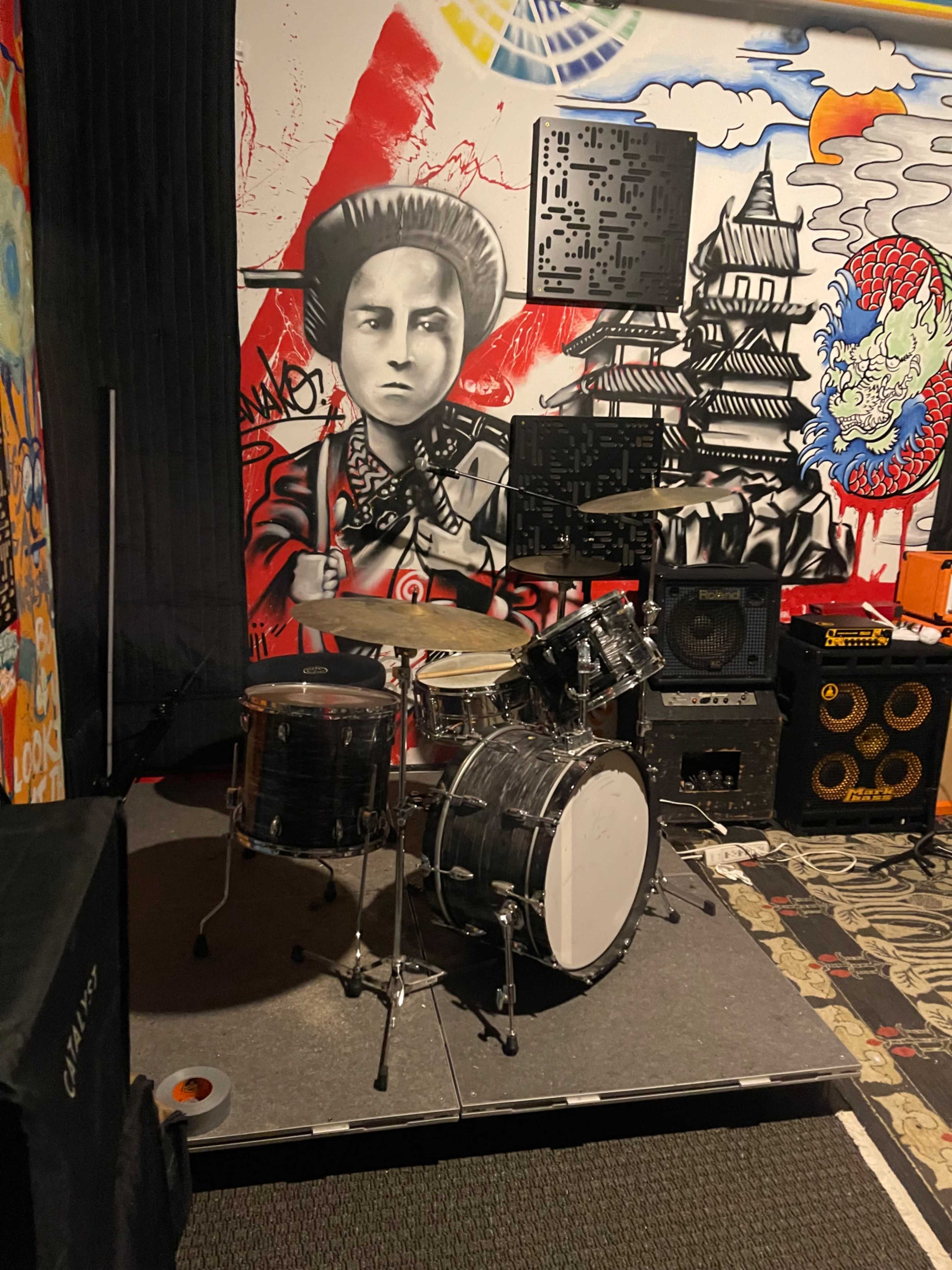 A drum set is positioned on a raised platform surrounded by vibrant graffiti artwork featuring a mural of an Asian figure and various abstract designs.