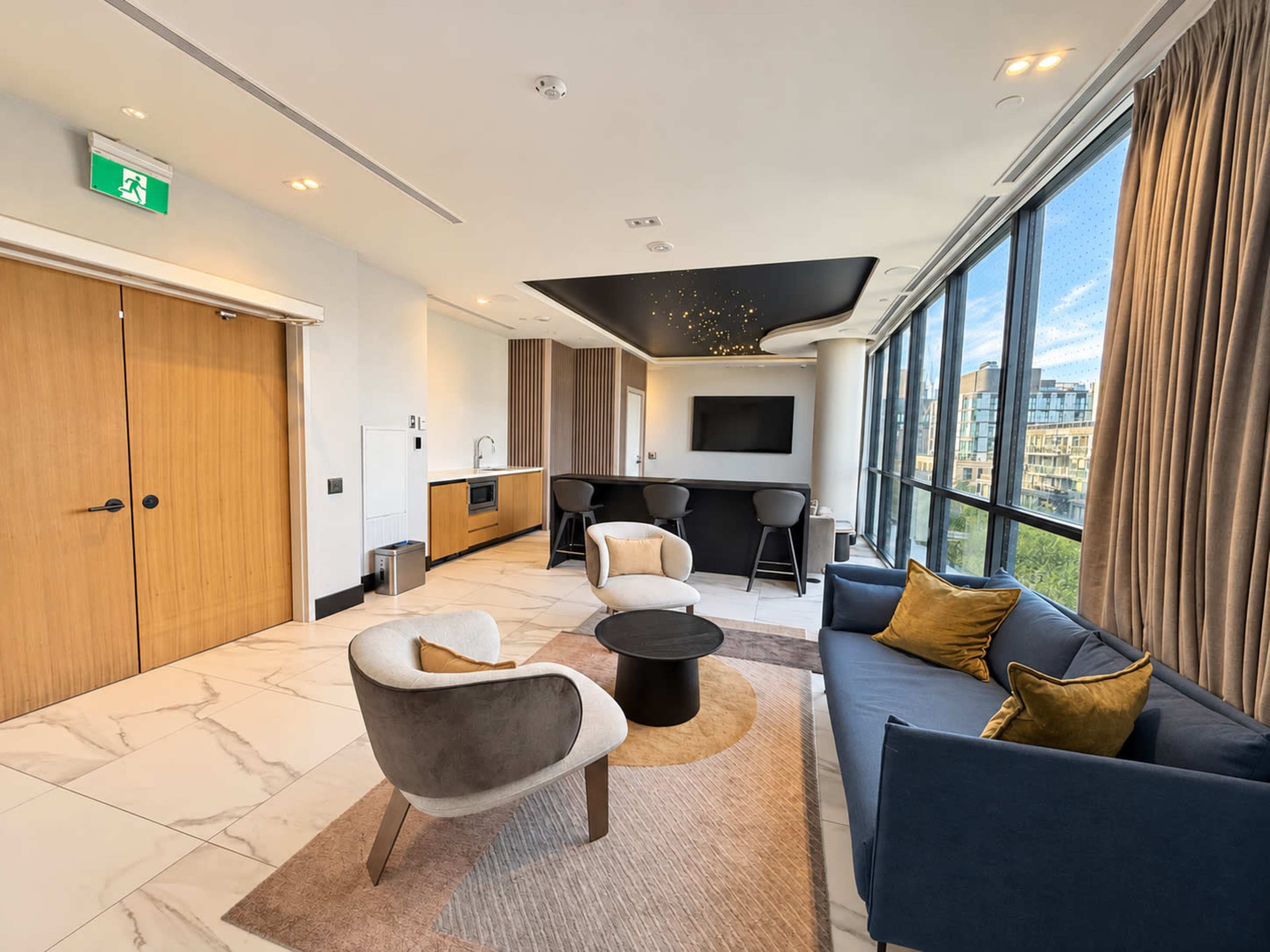 The image shows a modern living room with two armchairs, a sofa, a round coffee table, and large windows overlooking a cityscape.
