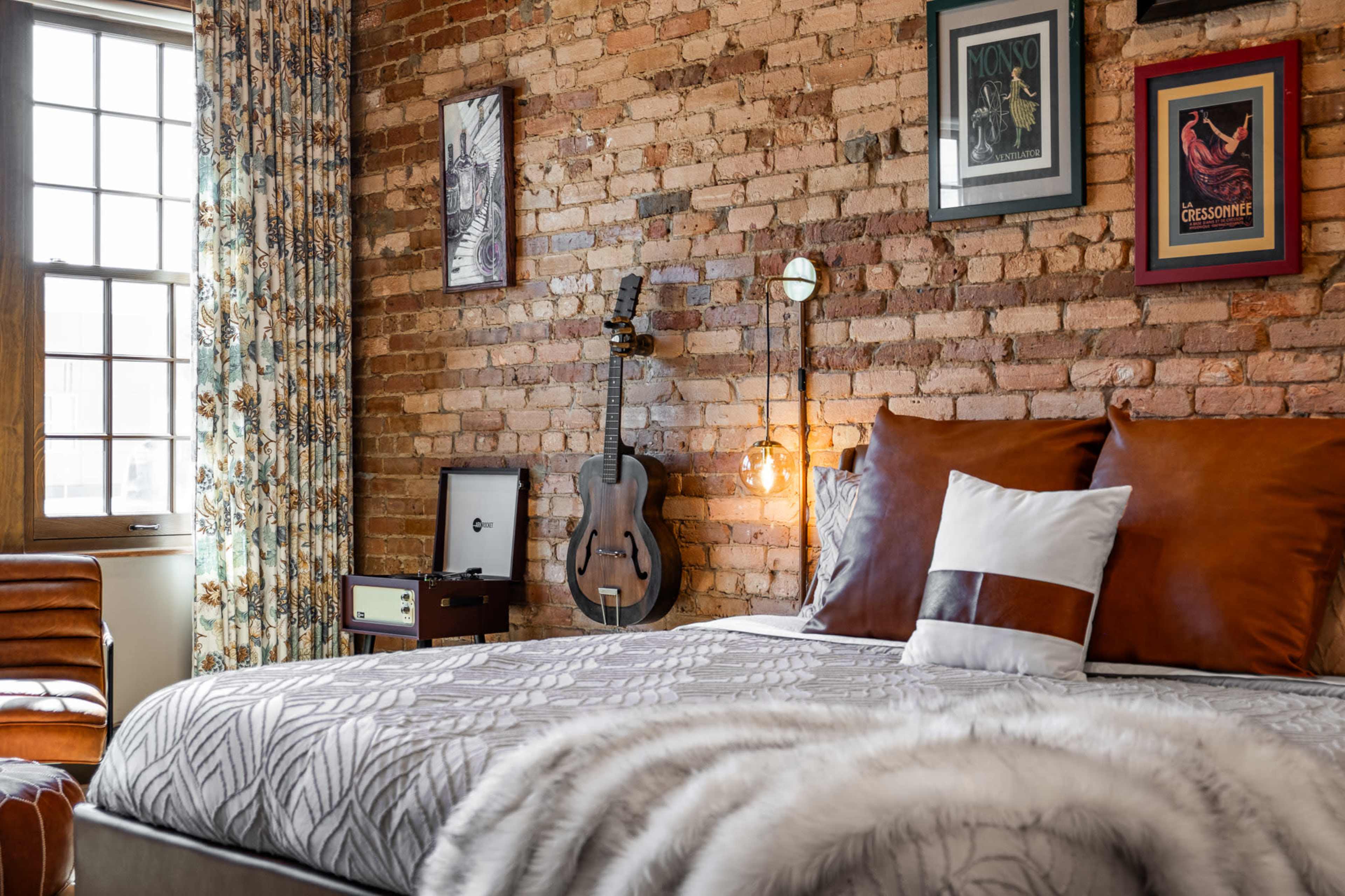 A stylish bedroom features a brick wall, a guitar hanging beside the window, and a large bed with layered bedding and decorative pillows.