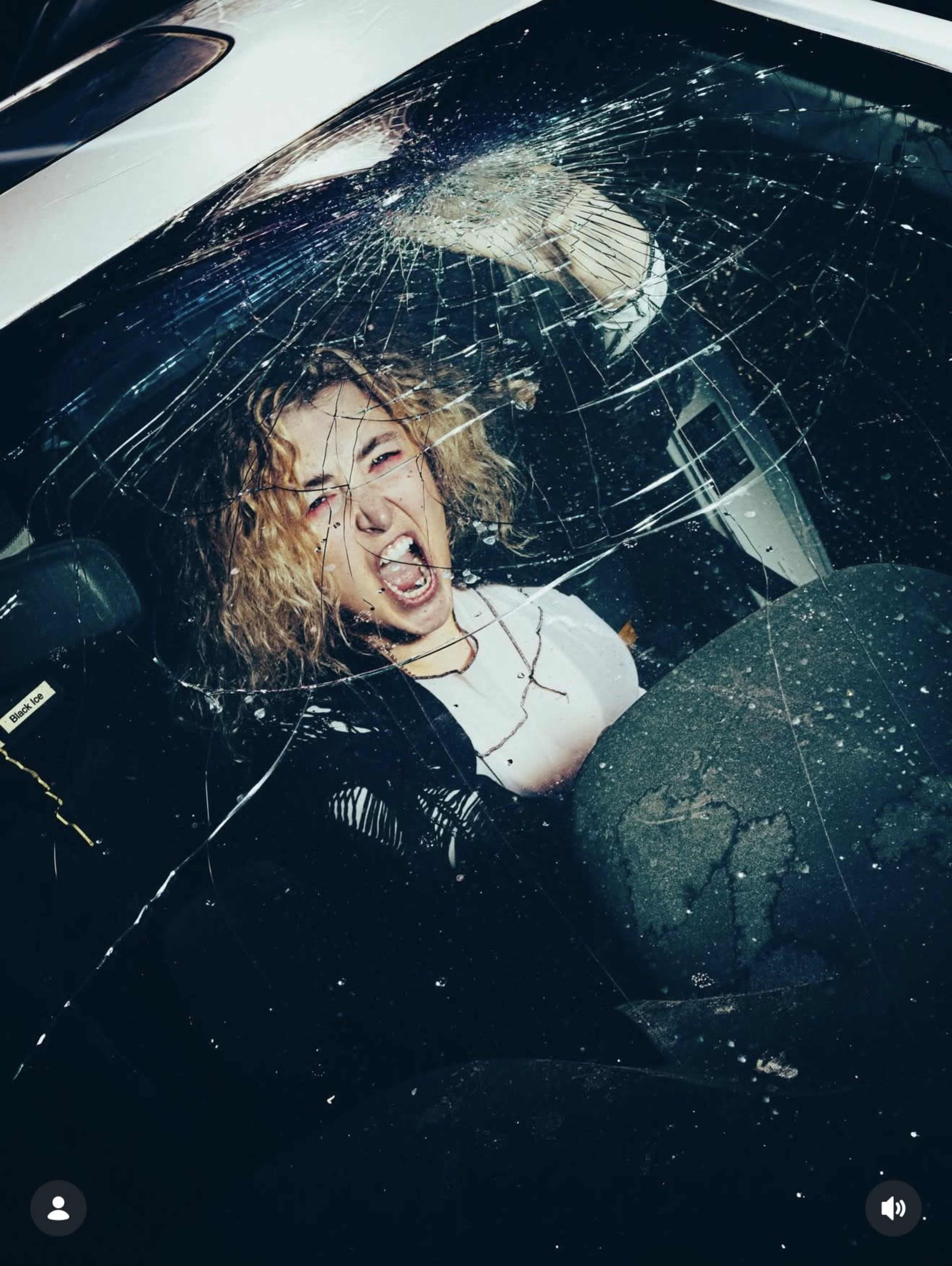 A woman with curly hair is seated inside a car, screaming while the windshield is shattered.