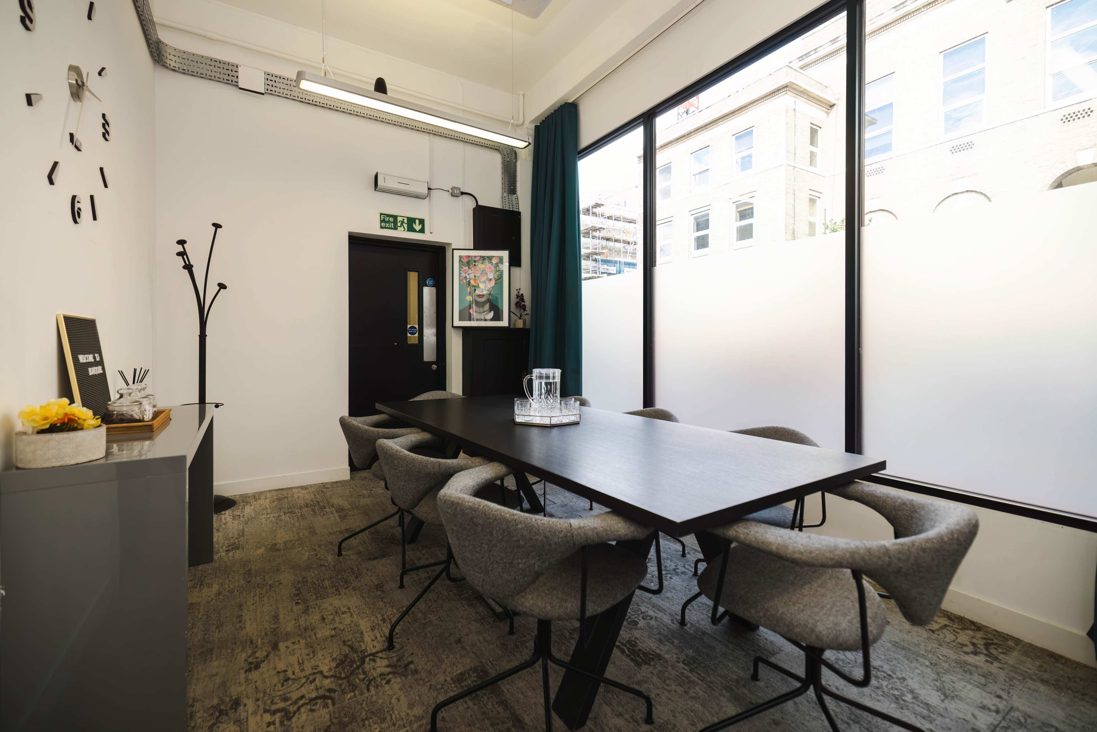 A meeting room features a long black table surrounded by gray fabric chairs and large windows that provide natural light.