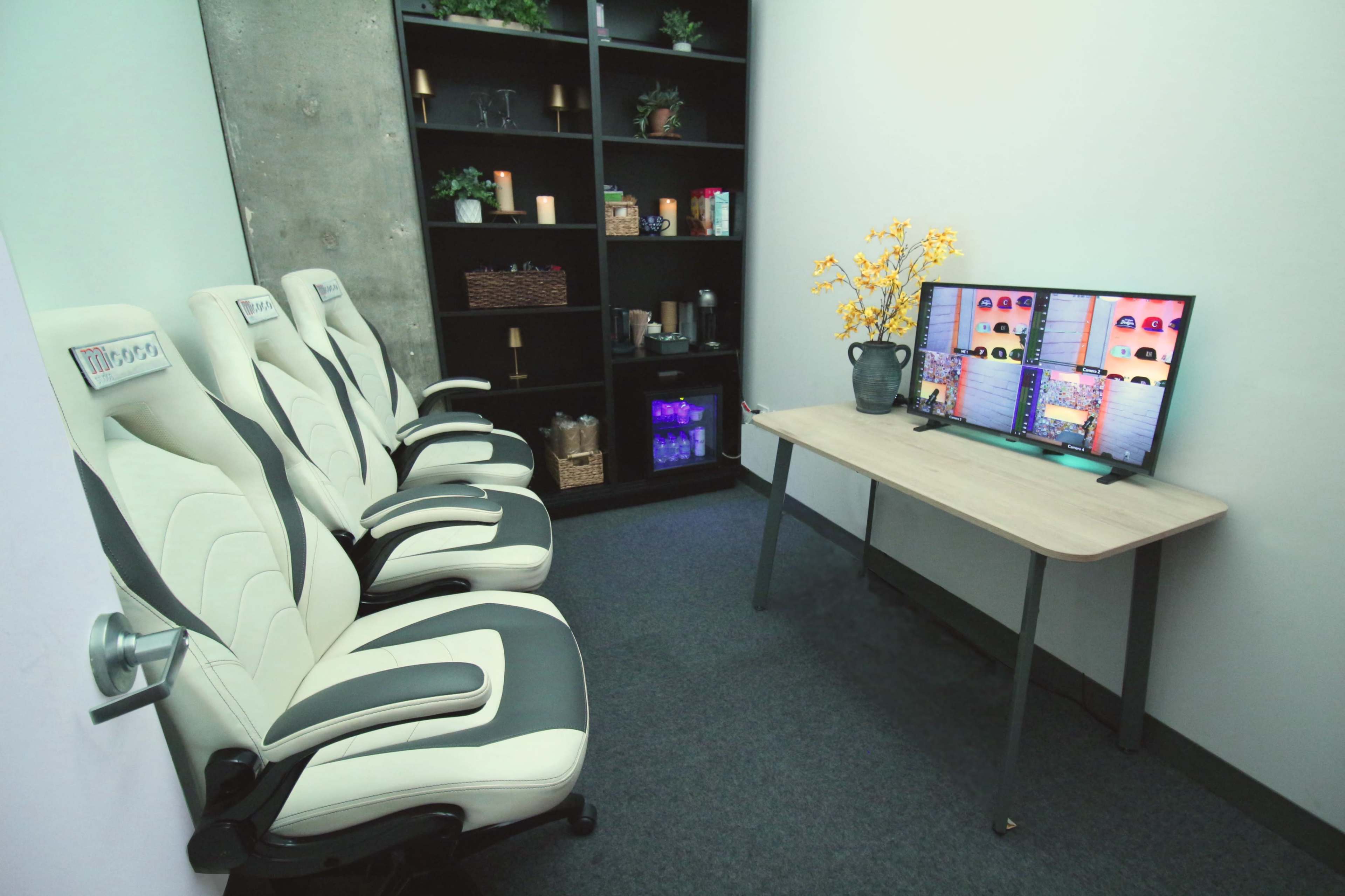 The image shows a small room with three ergonomic chairs, a wooden table, and a television displaying colorful images, next to a shelf filled with decorative items.