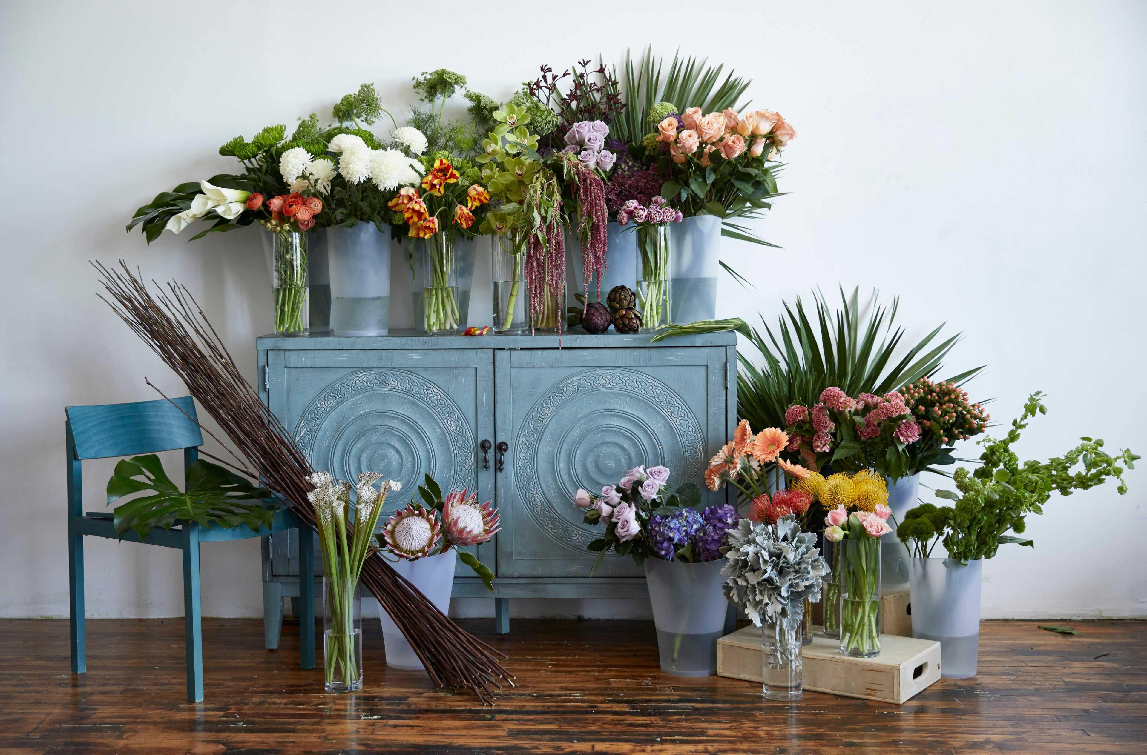 A blue cabinet surrounded by an array of colorful floral arrangements in vases, placed on the floor and a nearby chair.