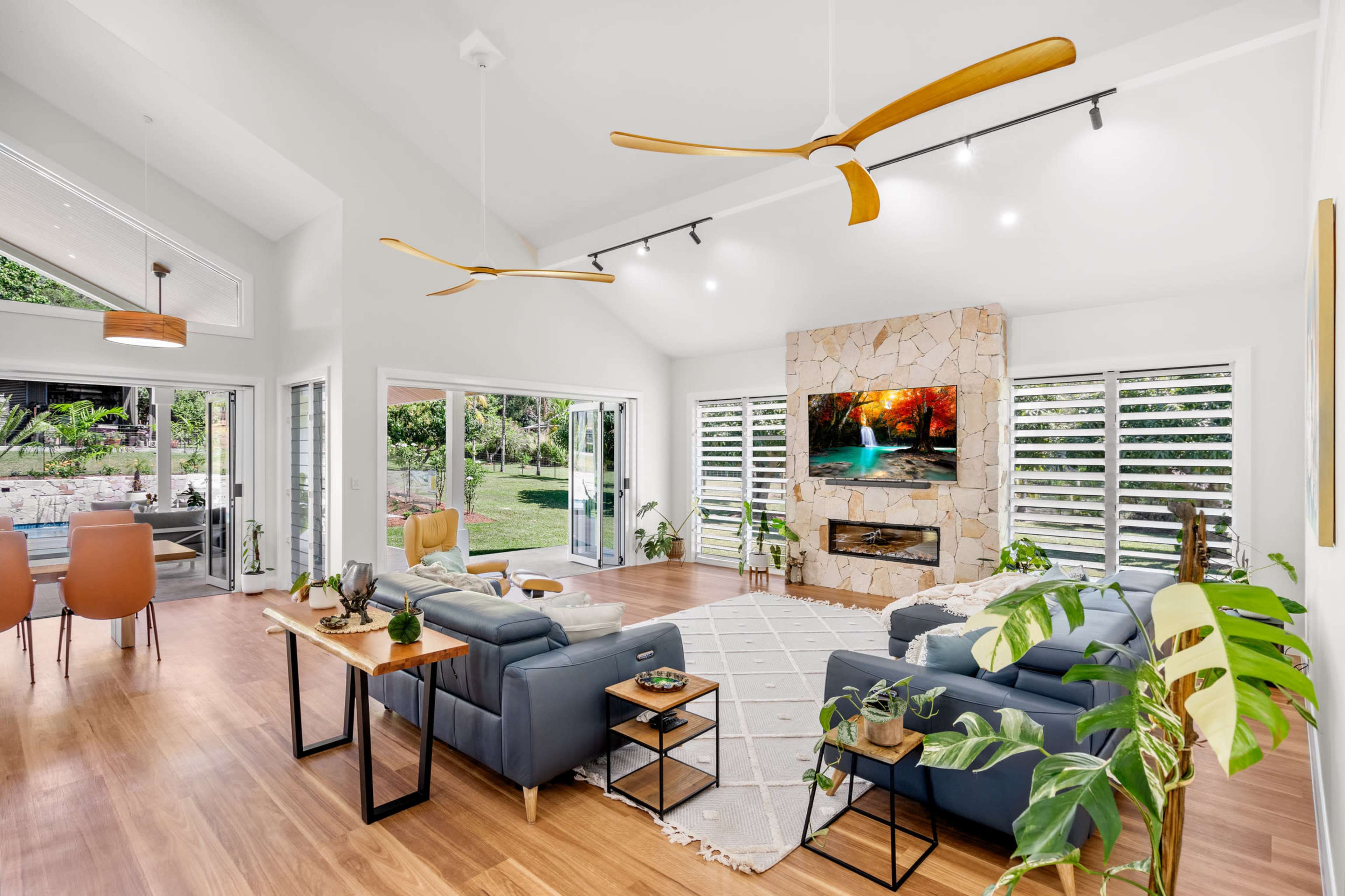 The image shows a modern living room with a stone fireplace, large windows, and a blend of comfortable seating and indoor plants.