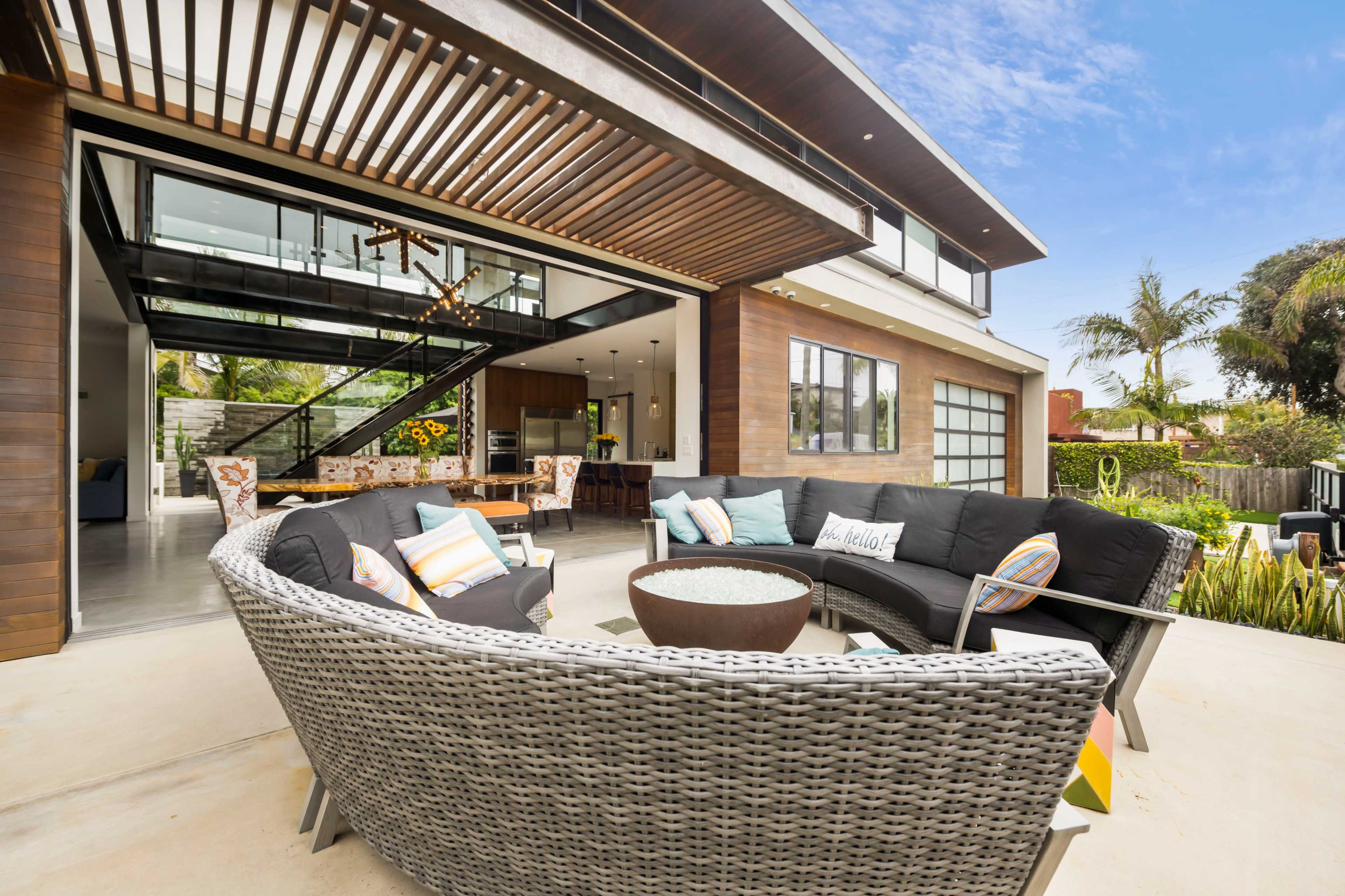 The image shows a modern outdoor seating area with a circular arrangement of wicker chairs surrounding a fire pit, adjacent to a spacious glass-fronted house.
