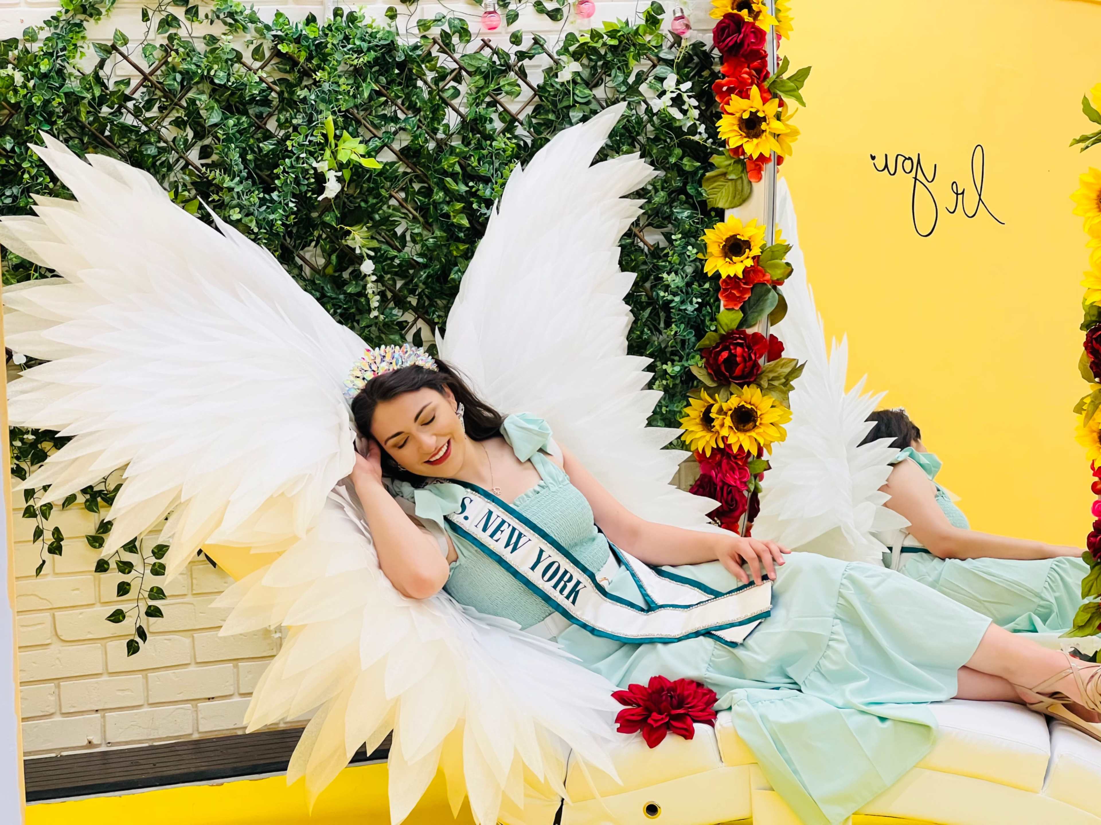 A woman wearing a sash labeled "Miss New York" reclines on a bench with large, decorative white wings surrounded by colorful floral accents against a bright yellow background.