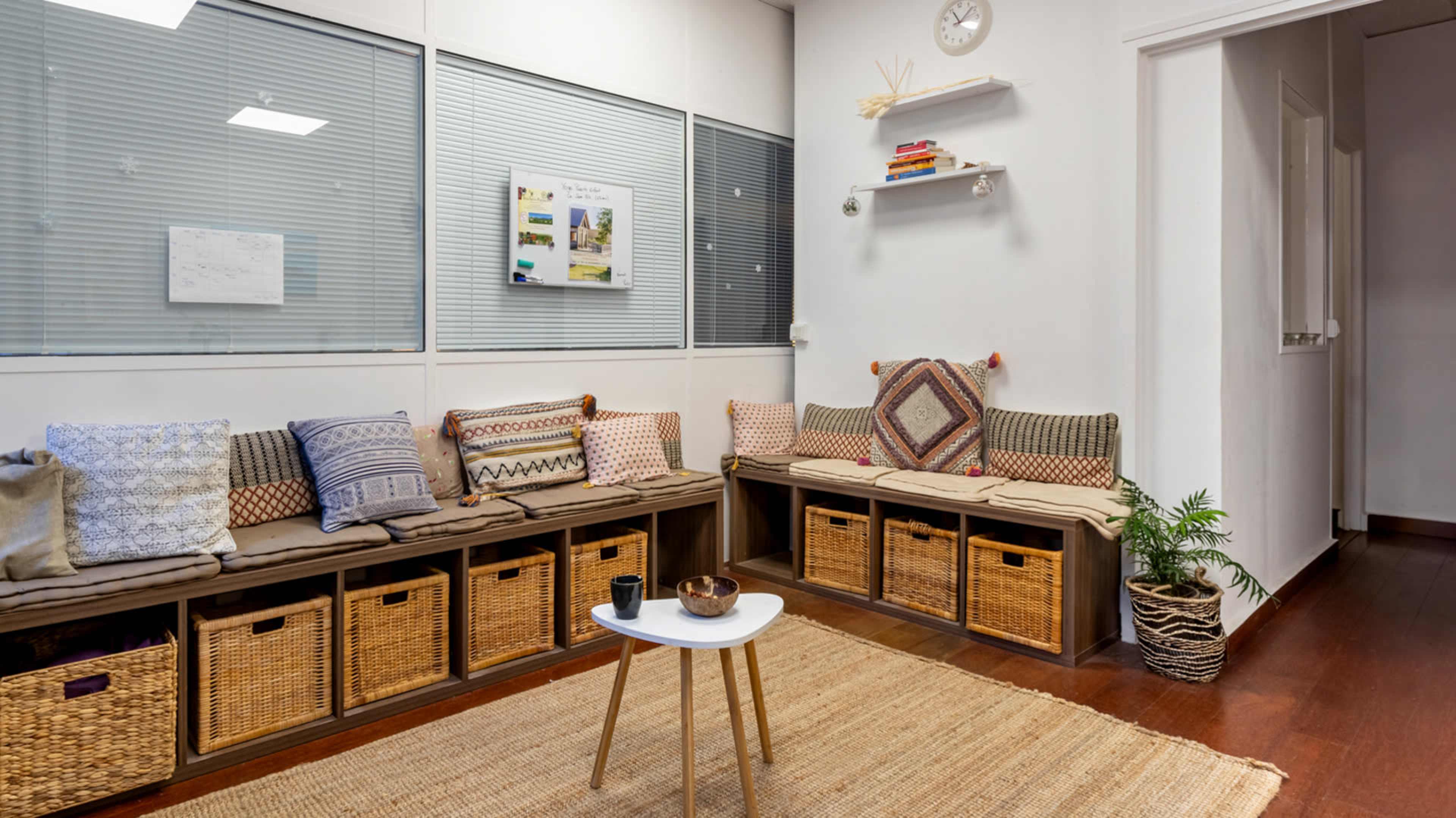 The image shows a cozy waiting area with a long bench covered in patterned cushions, storage baskets underneath, and a small table on a woven rug.