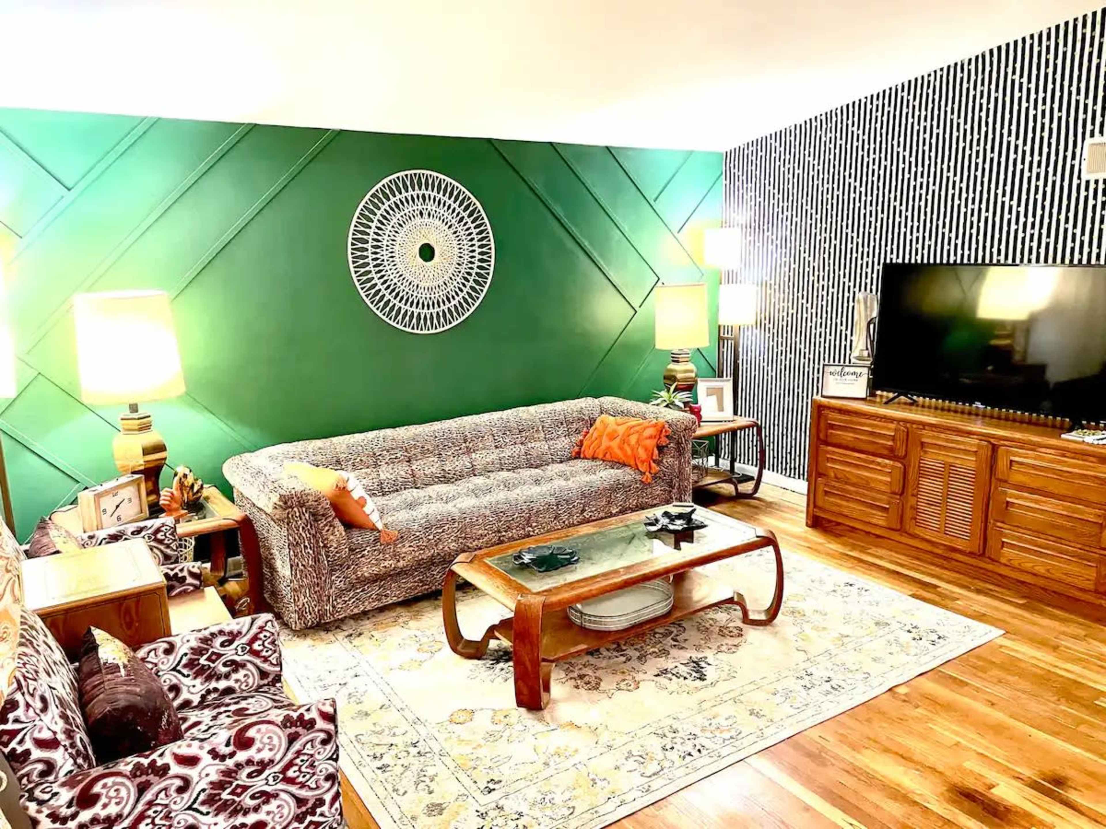 A living room featuring a gray patterned sofa, a green accent wall with a decorative circular piece, and a television on a wooden stand against a striped wall.