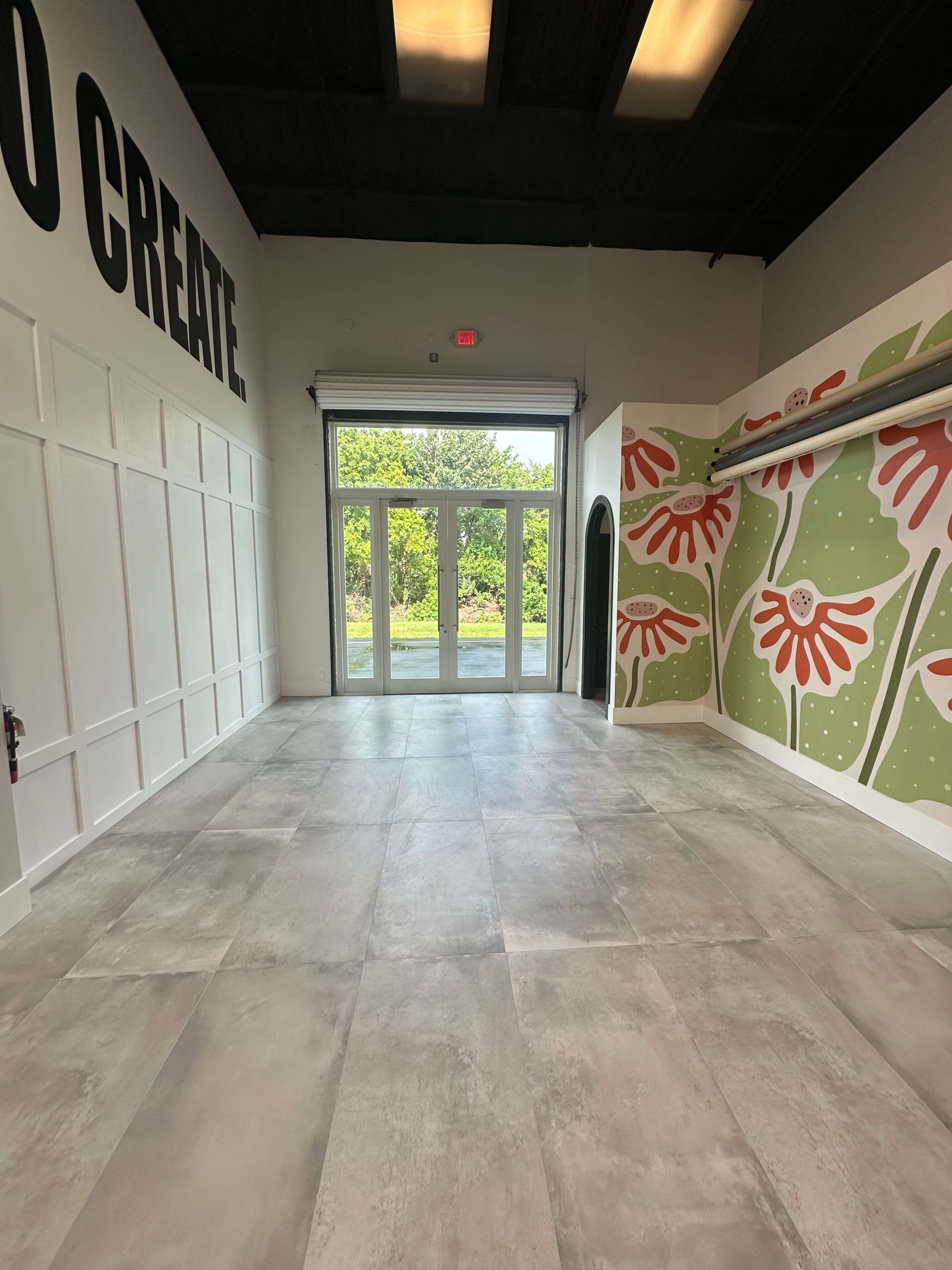 The interior of a spacious, empty room features large windows opening to a green area and a mural with floral designs on one wall.