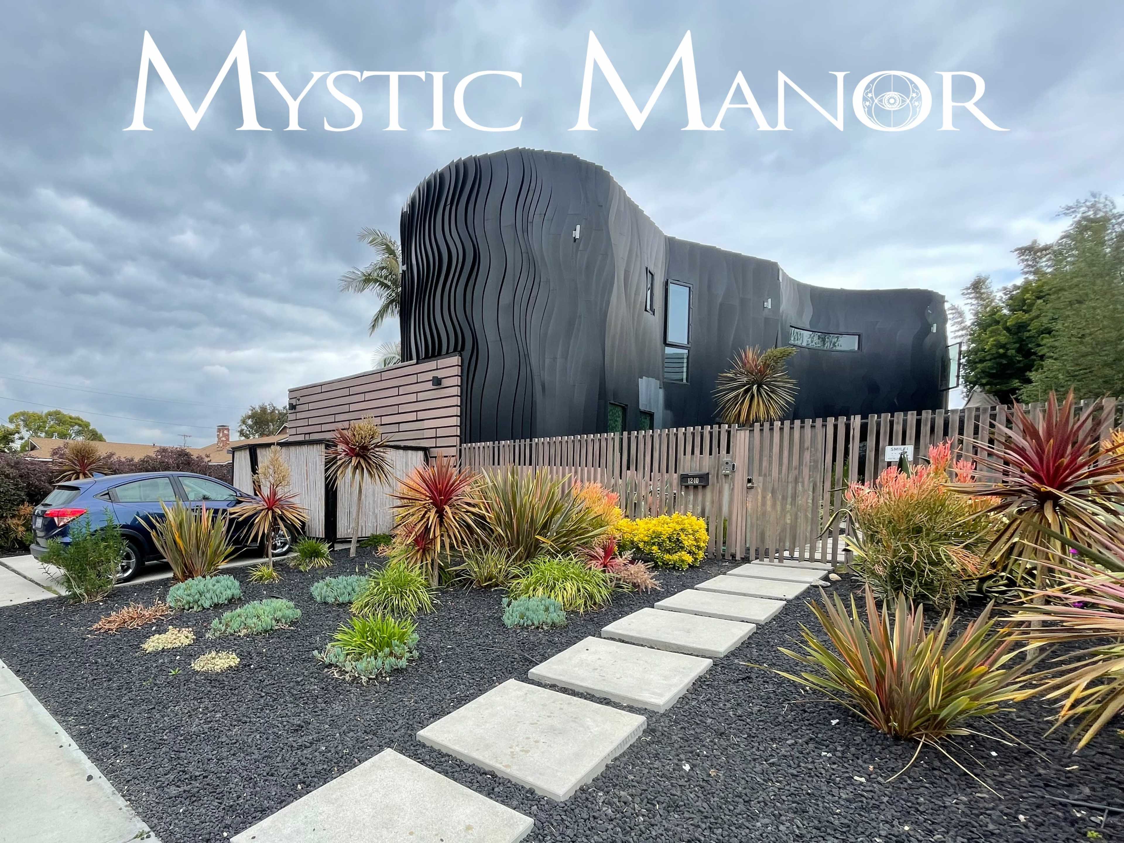 A modern black building with a wavy exterior is set behind a wooden fence, surrounded by landscaped gardens and a stone pathway.