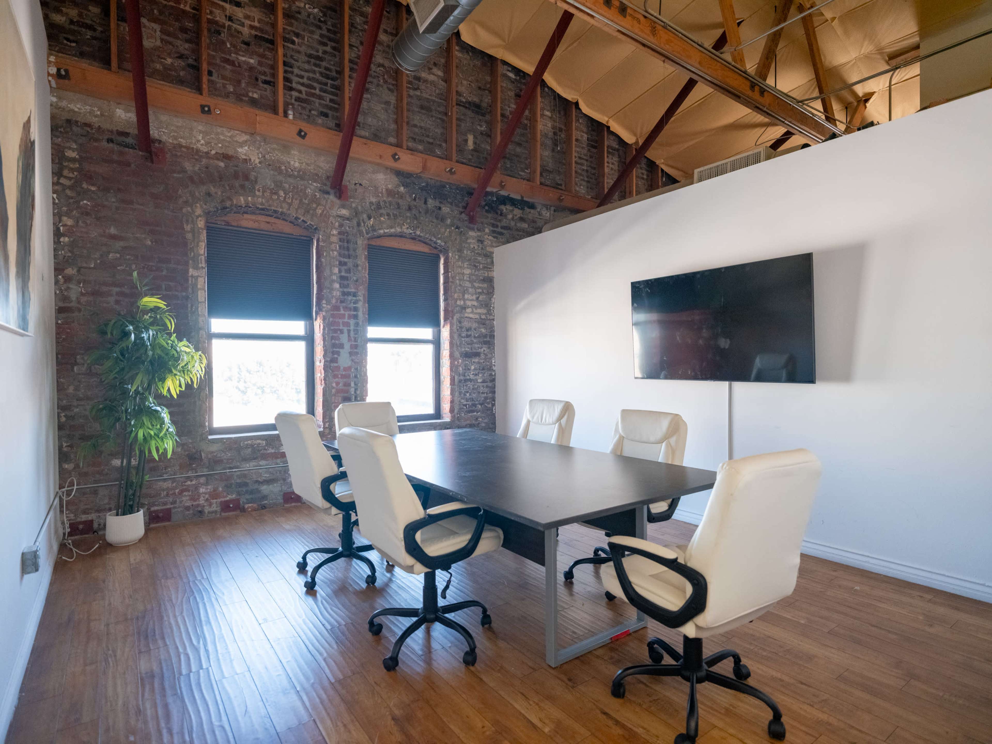 A modern conference room features a large table surrounded by six white office chairs and a mounted television on the wall, with exposed brick walls and large windows.