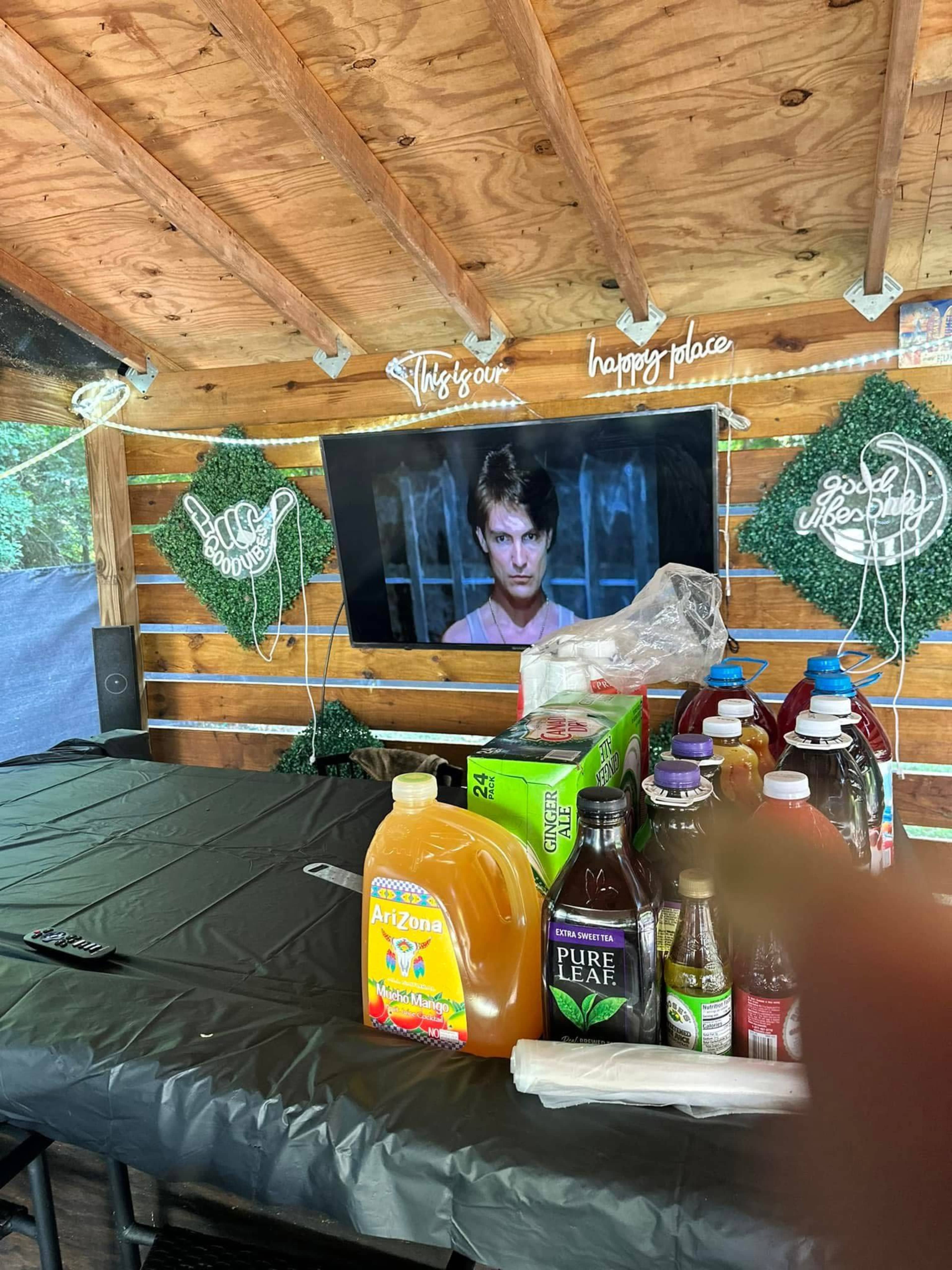 The image shows a wooden outdoor space with a black table, various drink containers, and a mounted television displaying a scene from a film.
