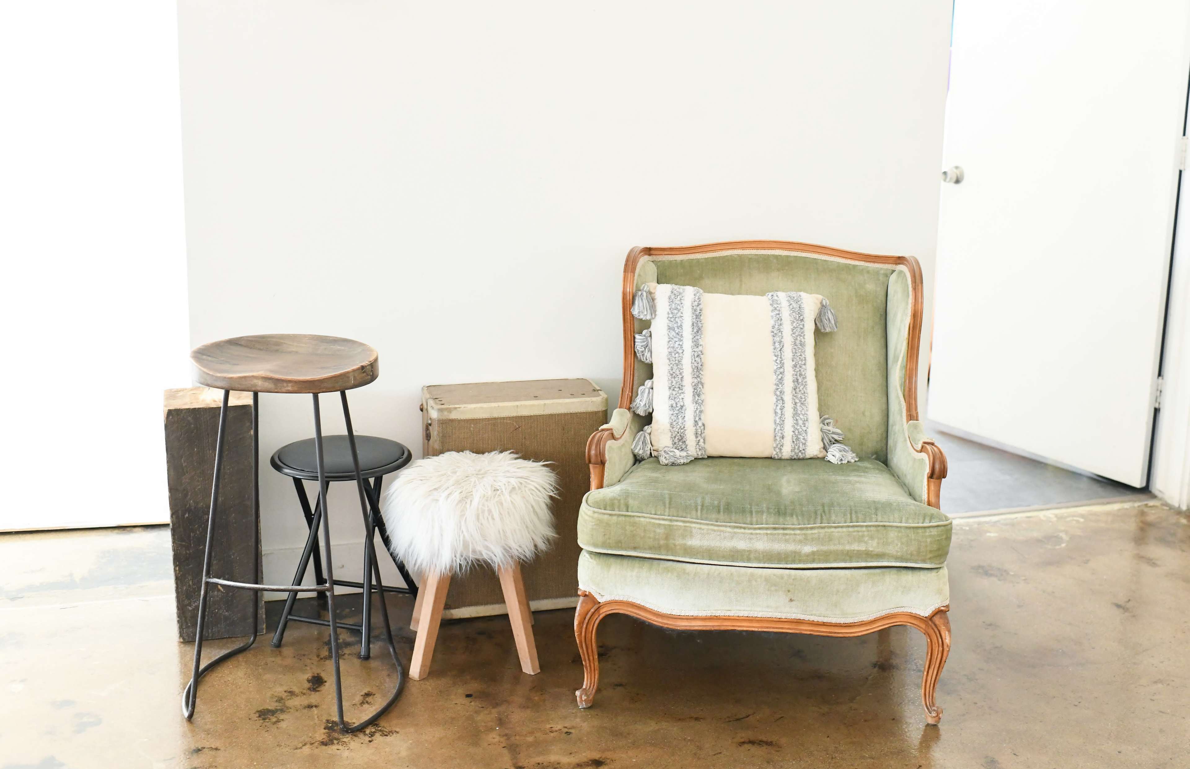 A green upholstered armchair with decorative pillows is positioned next to a small wooden stool and a fluffy white ottoman, alongside a wooden crate in a minimalist indoor setting.