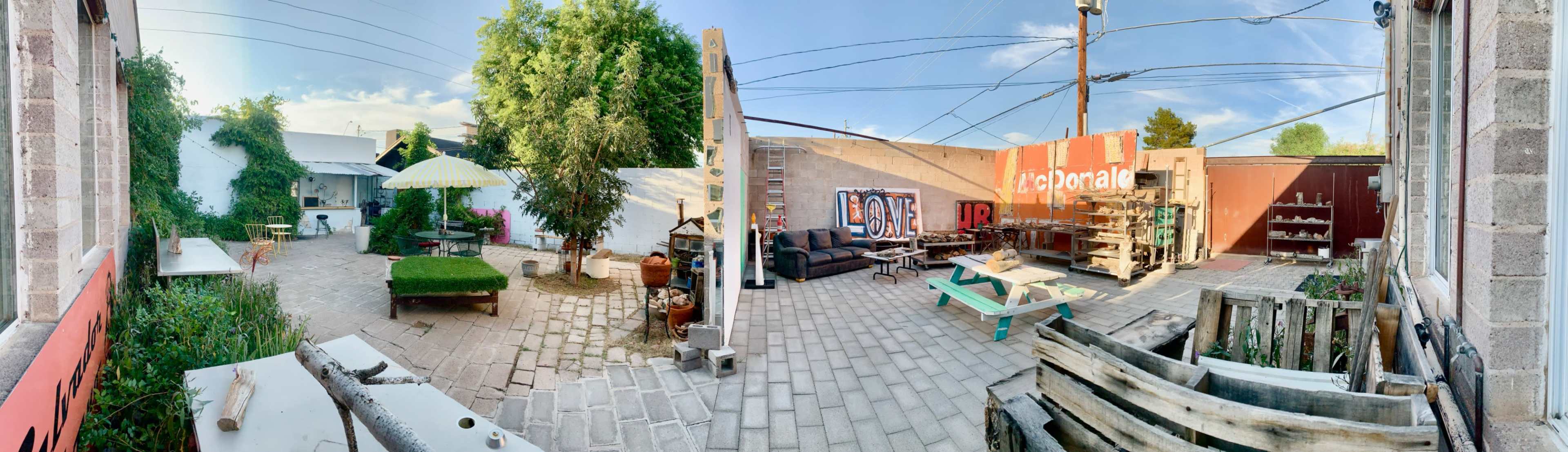 The image shows a panoramic view of a versatile outdoor space featuring a grassy area, seating arrangements, and decorative elements, surrounded by walls and power lines.