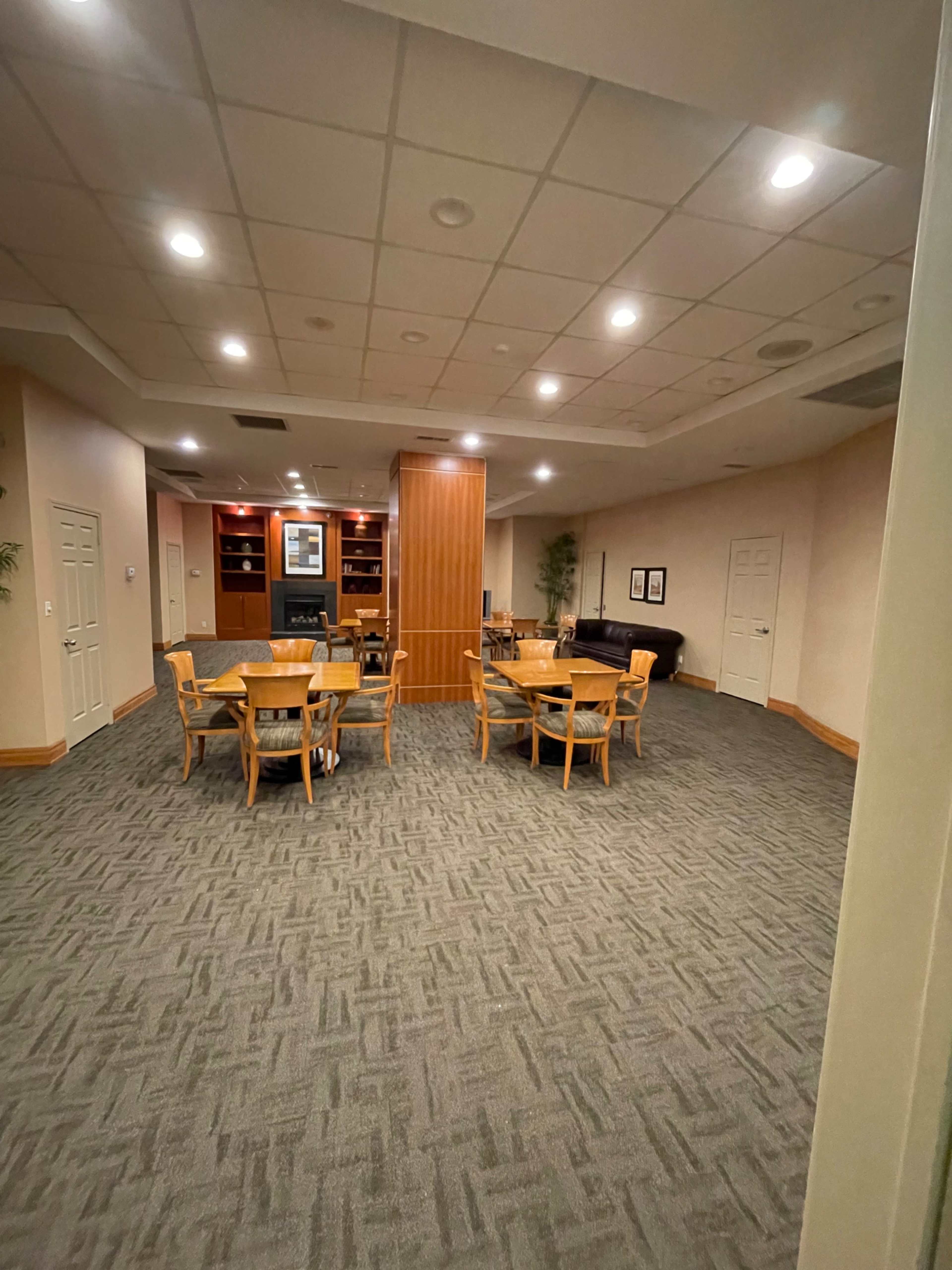 The image shows a well-lit, empty room featuring several wooden tables and chairs arranged around a central area.