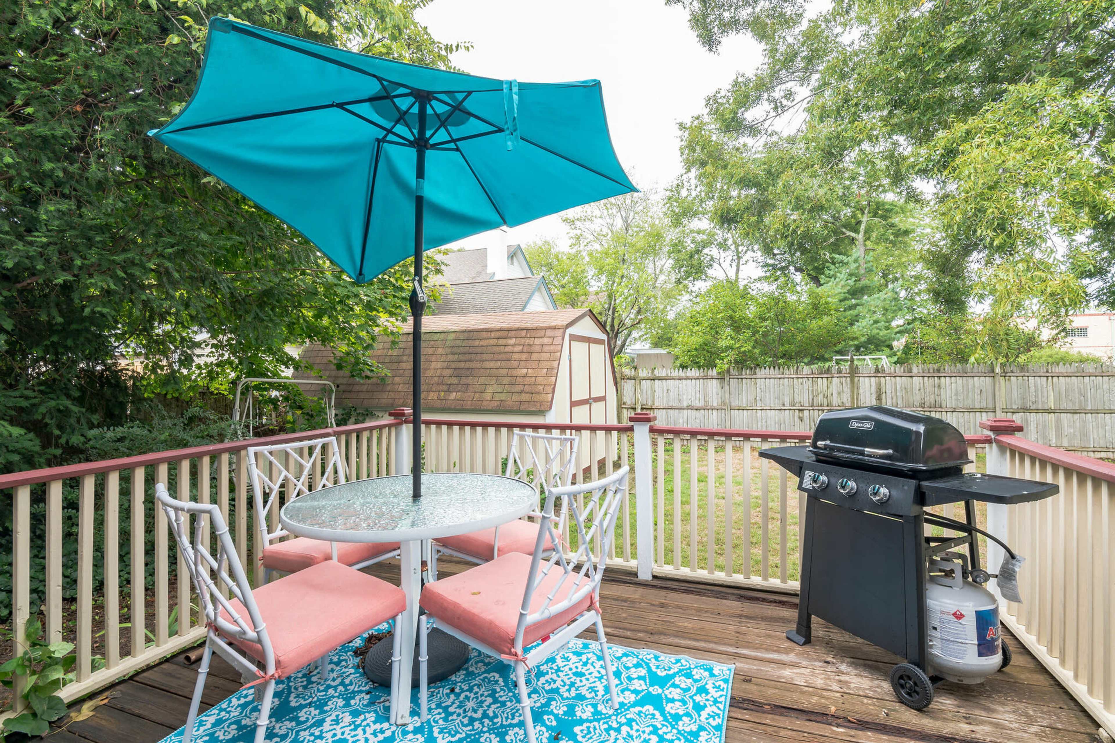 A deck features a round table with pink chairs, an umbrella, and a grill, surrounded by greenery and a shed in the background.