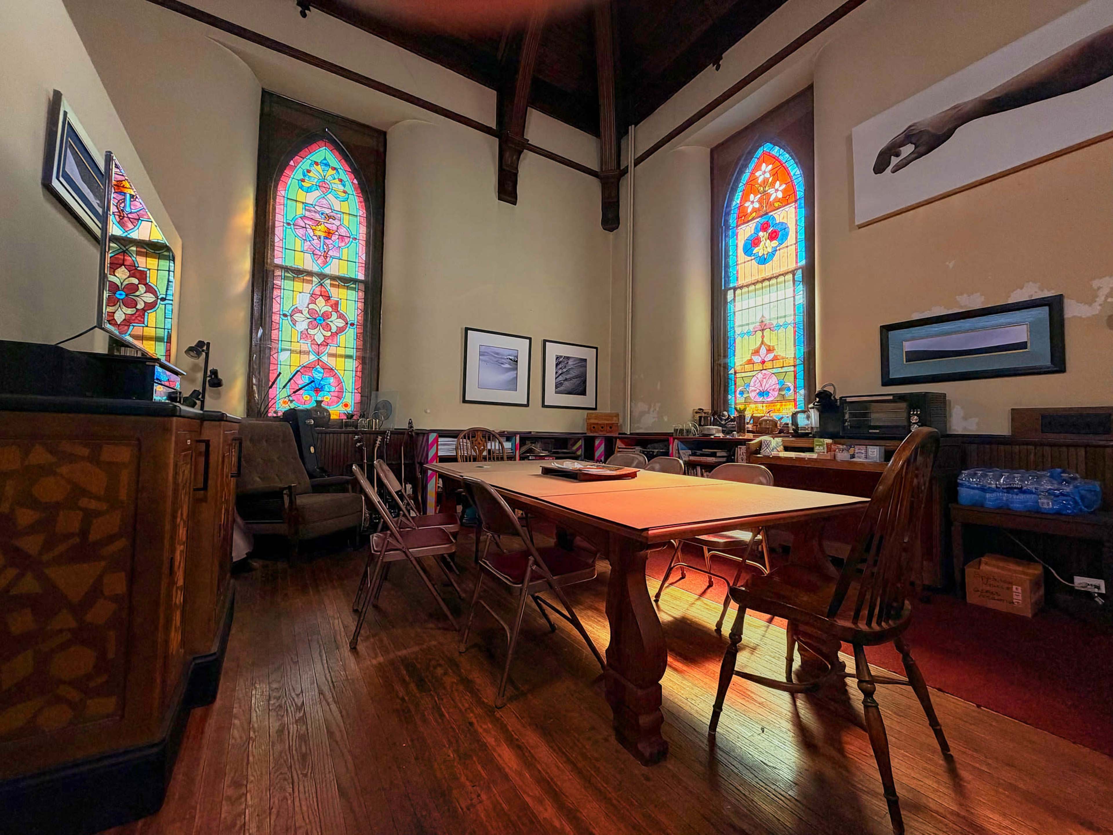 The image shows a sunlit room with stained glass windows, a large table surrounded by chairs, and various framed artworks on the walls.