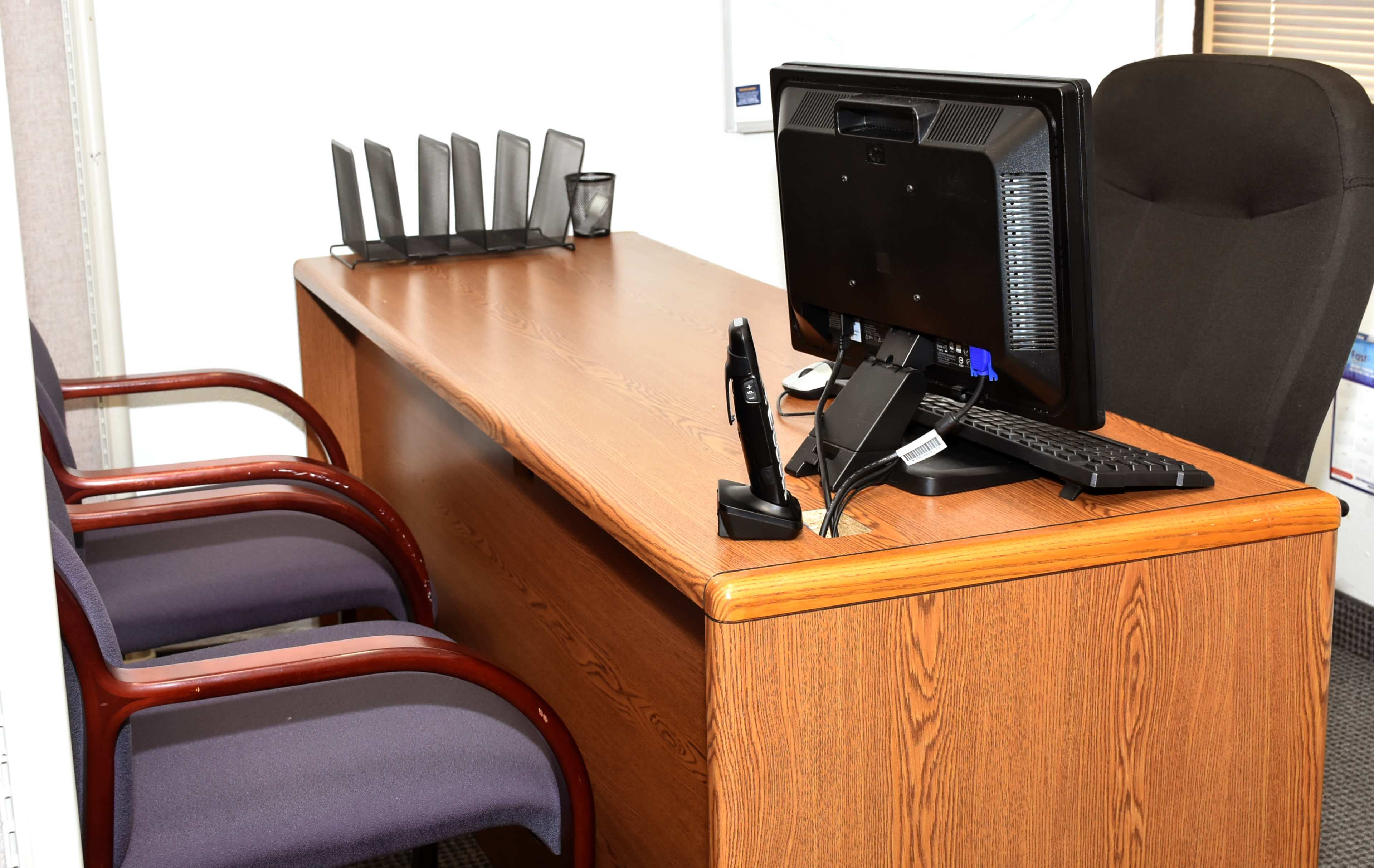 A simple wooden desk with a computer monitor, a wireless keyboard, and two office chairs positioned in front.
