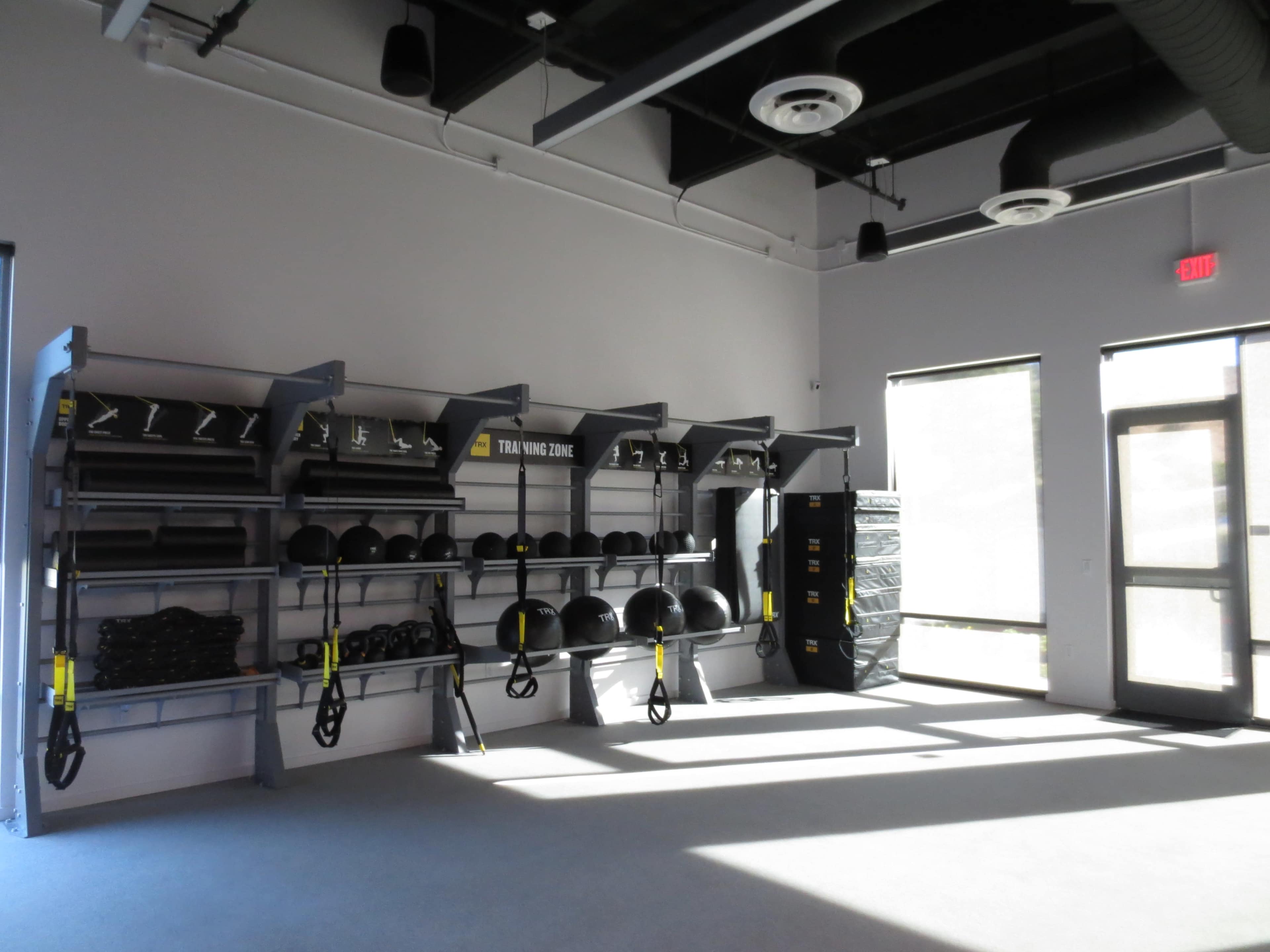 The image shows a fitness training area with wall-mounted shelves holding various exercise equipment, including weights and suspension trainers, along with large windows allowing natural light.