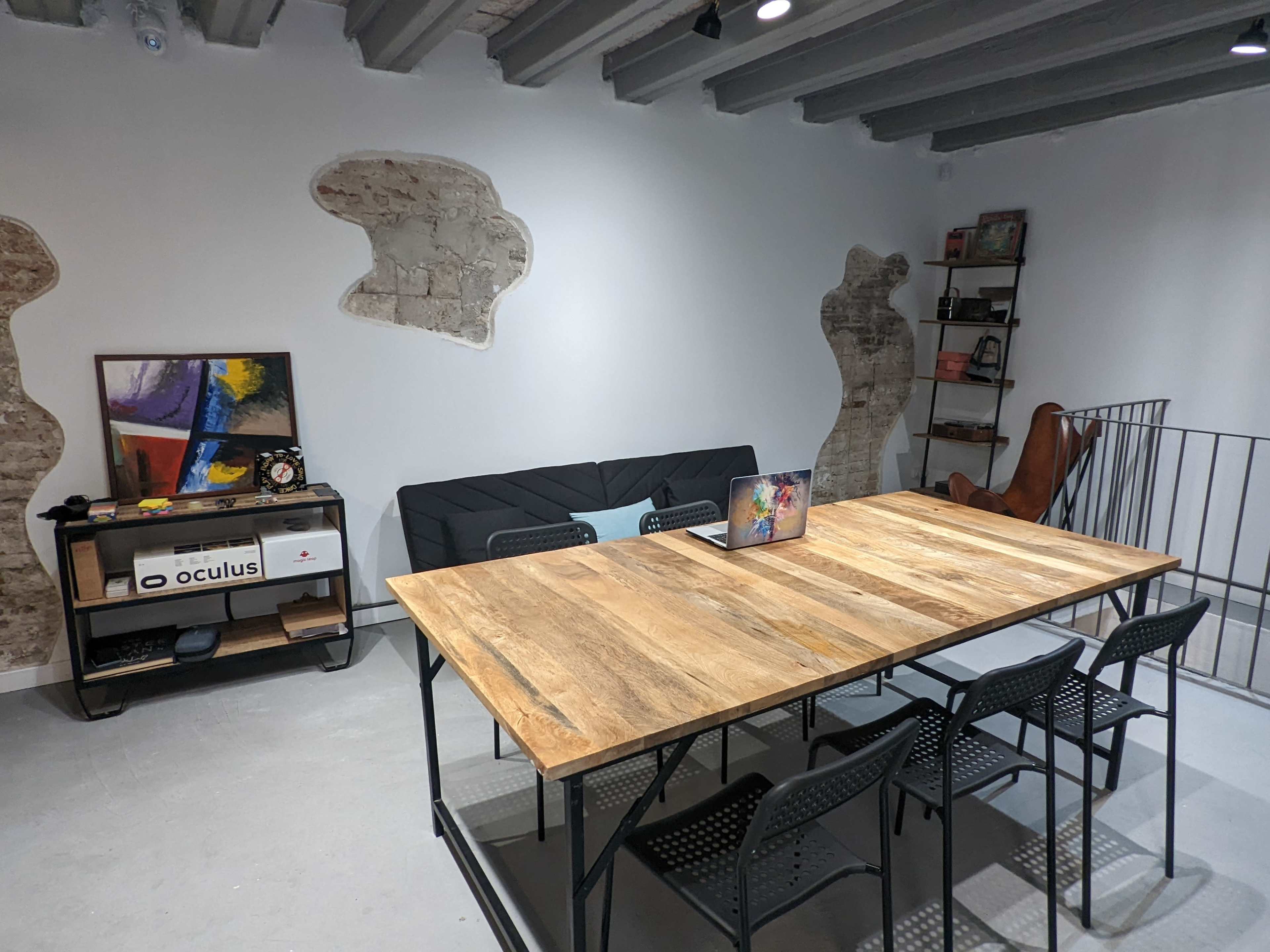The image shows a modern meeting room featuring a large wooden table with black chairs, a couch, a shelf with books, and an abstract wall design.