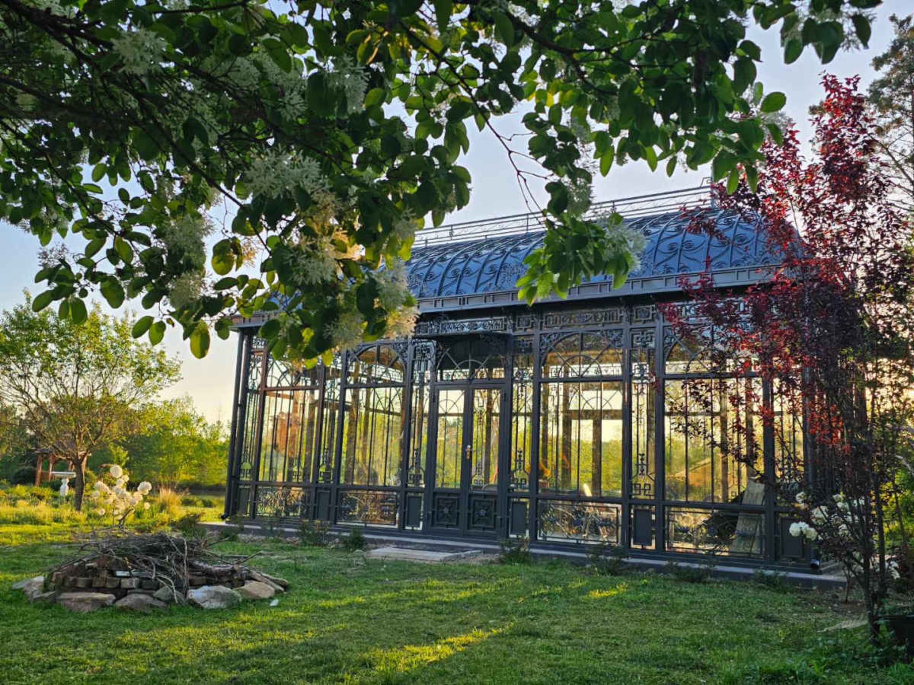 A metal-framed greenhouse surrounded by greenery and a stone arrangement in a garden.