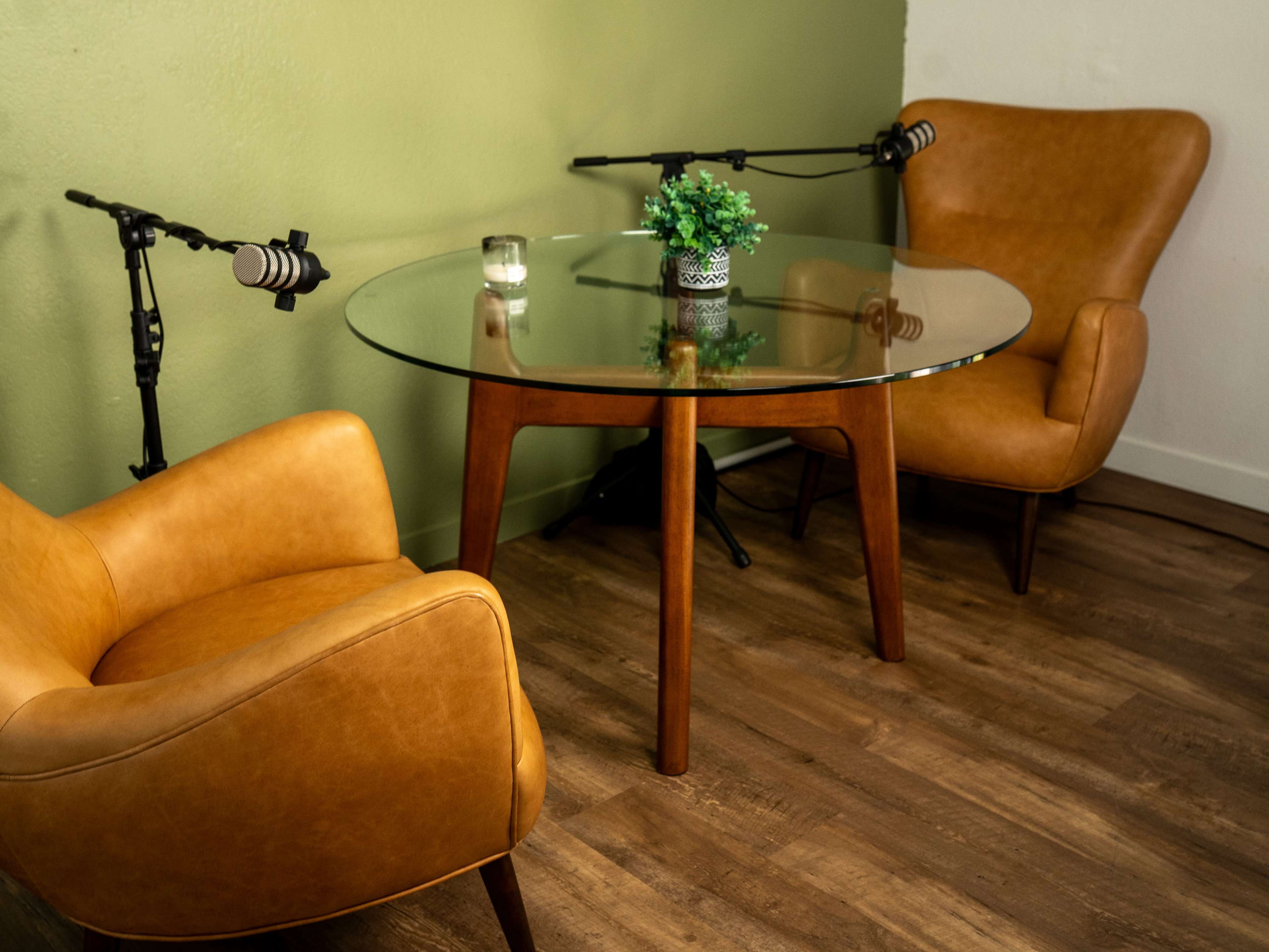 The image shows a small meeting space with a glass table and two brown leather armchairs, accompanied by microphones positioned nearby.