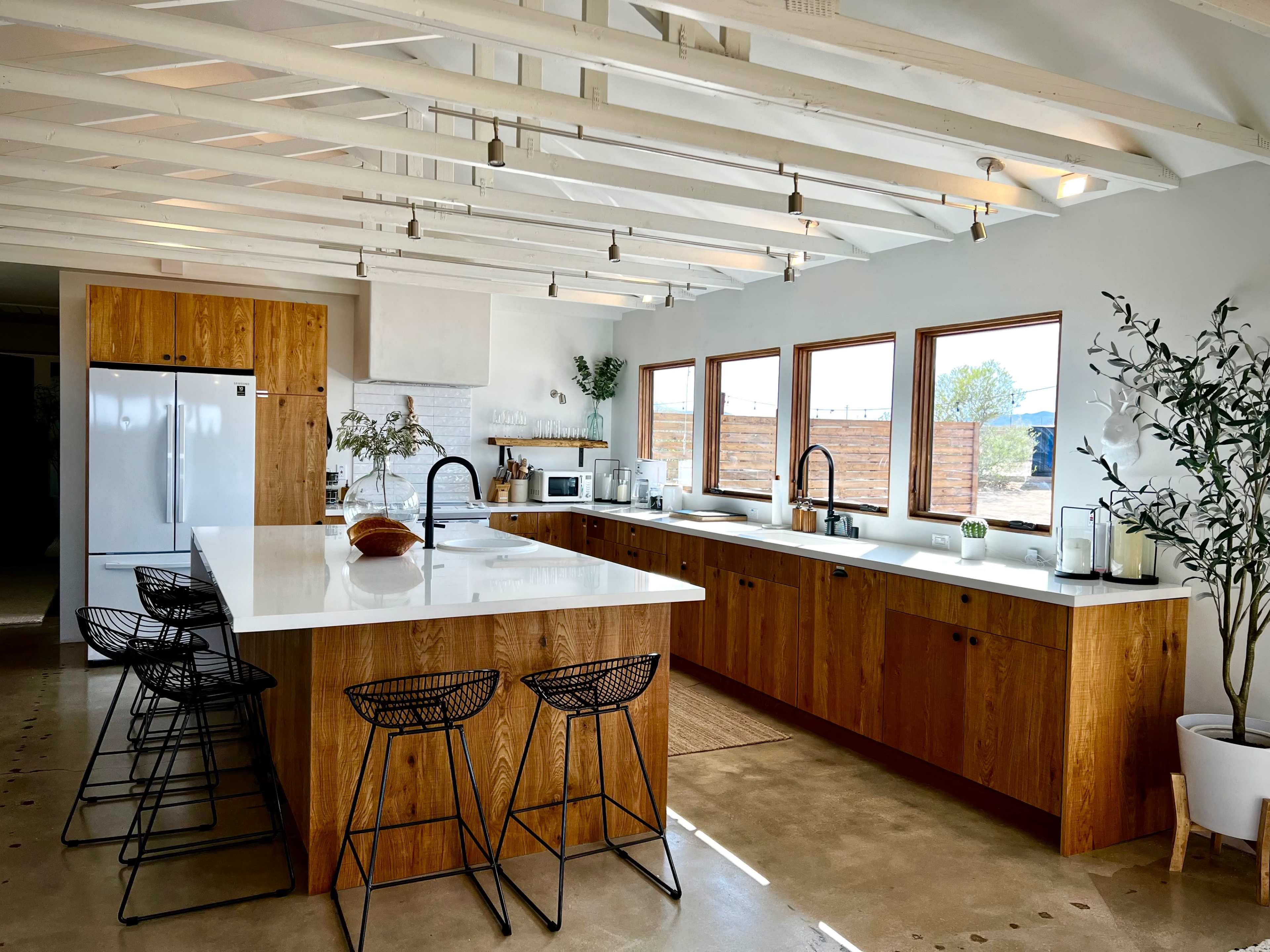 A modern kitchen features a large island with four black bar stools, wooden cabinetry, and large windows providing natural light.