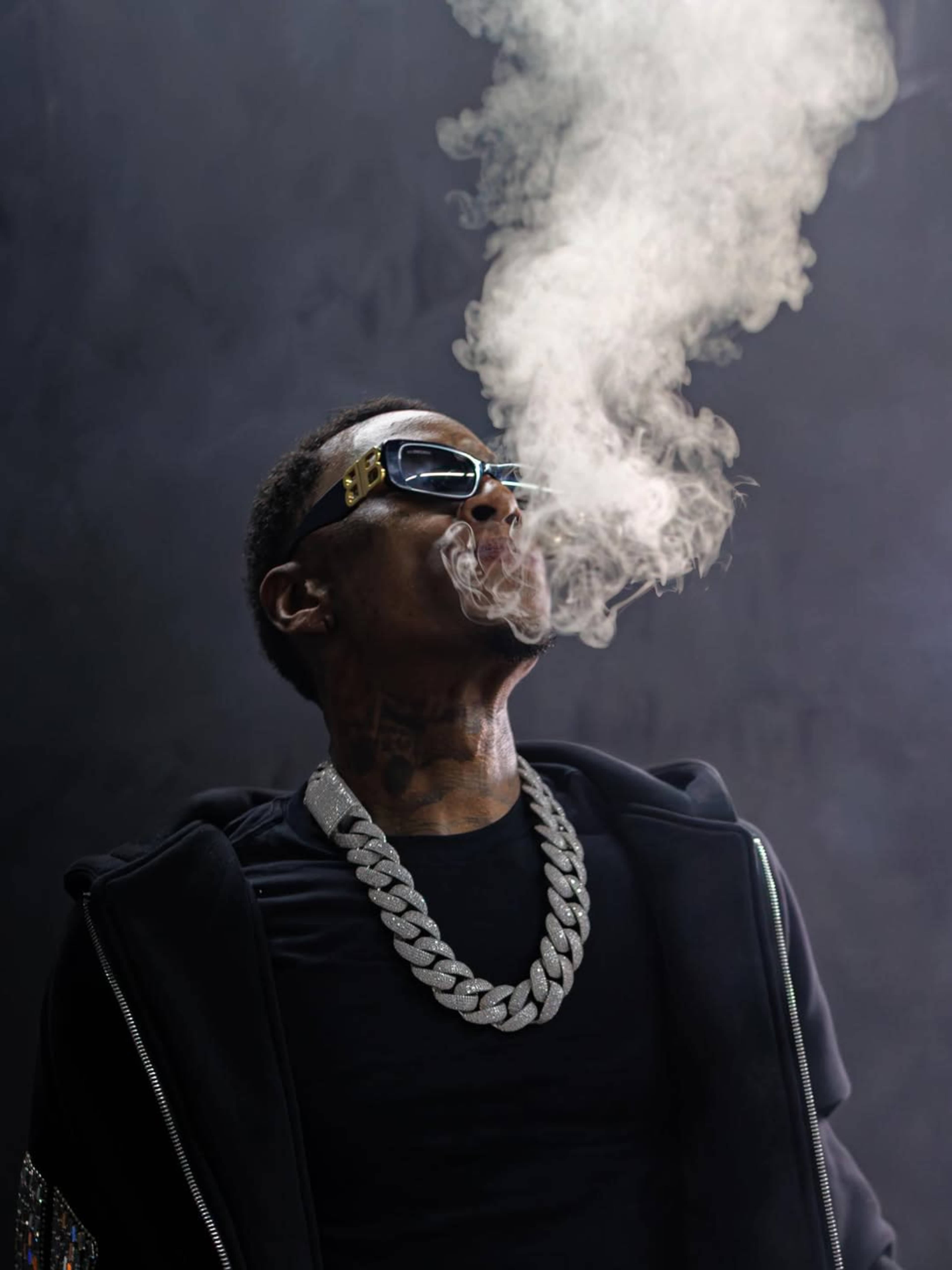 A person wearing sunglasses and a large chain exhales a cloud of smoke against a dark background.