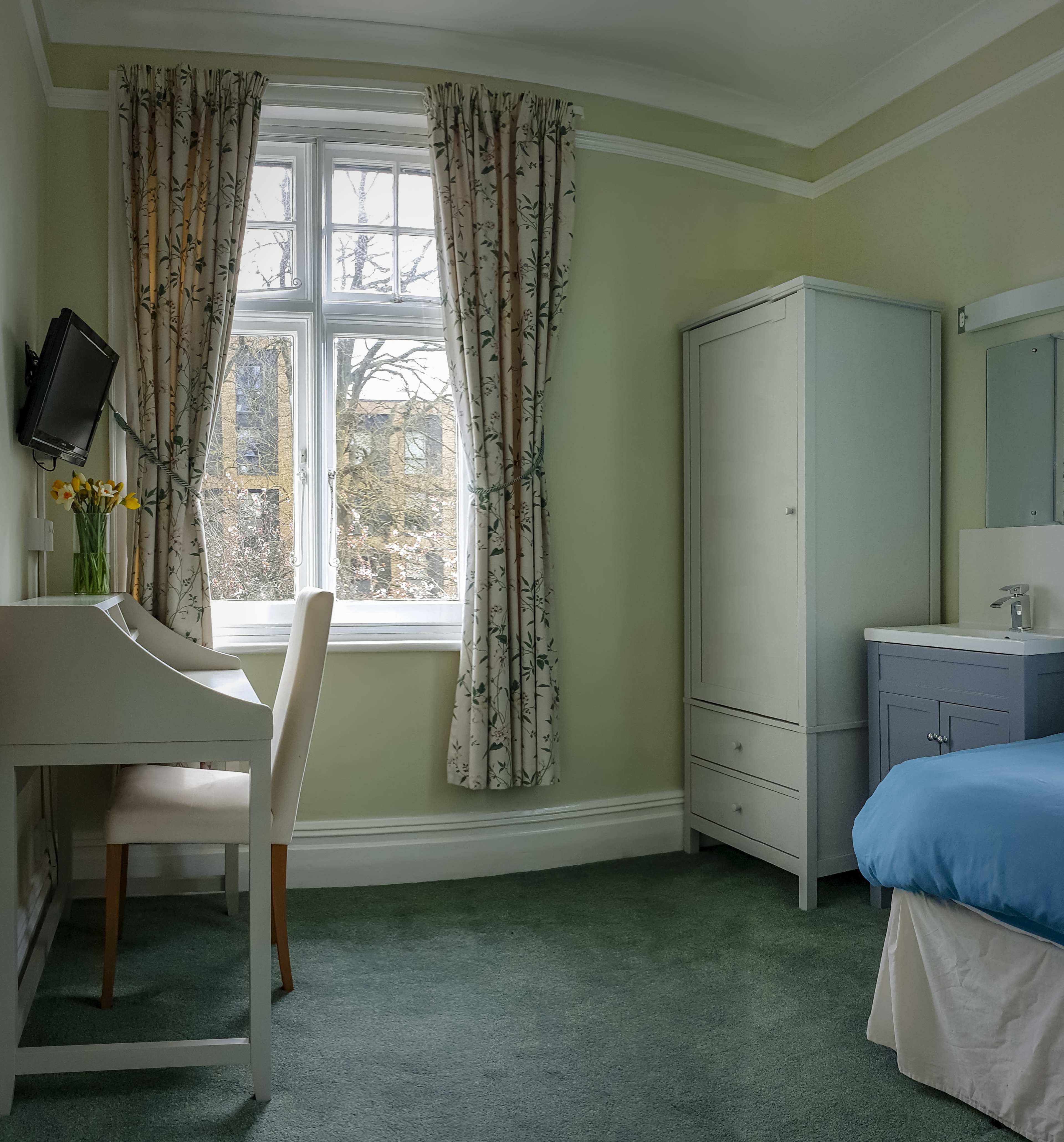 A tidy bedroom featuring a bed, a small desk with a chair, a wardrobe, and a window with patterned curtains.