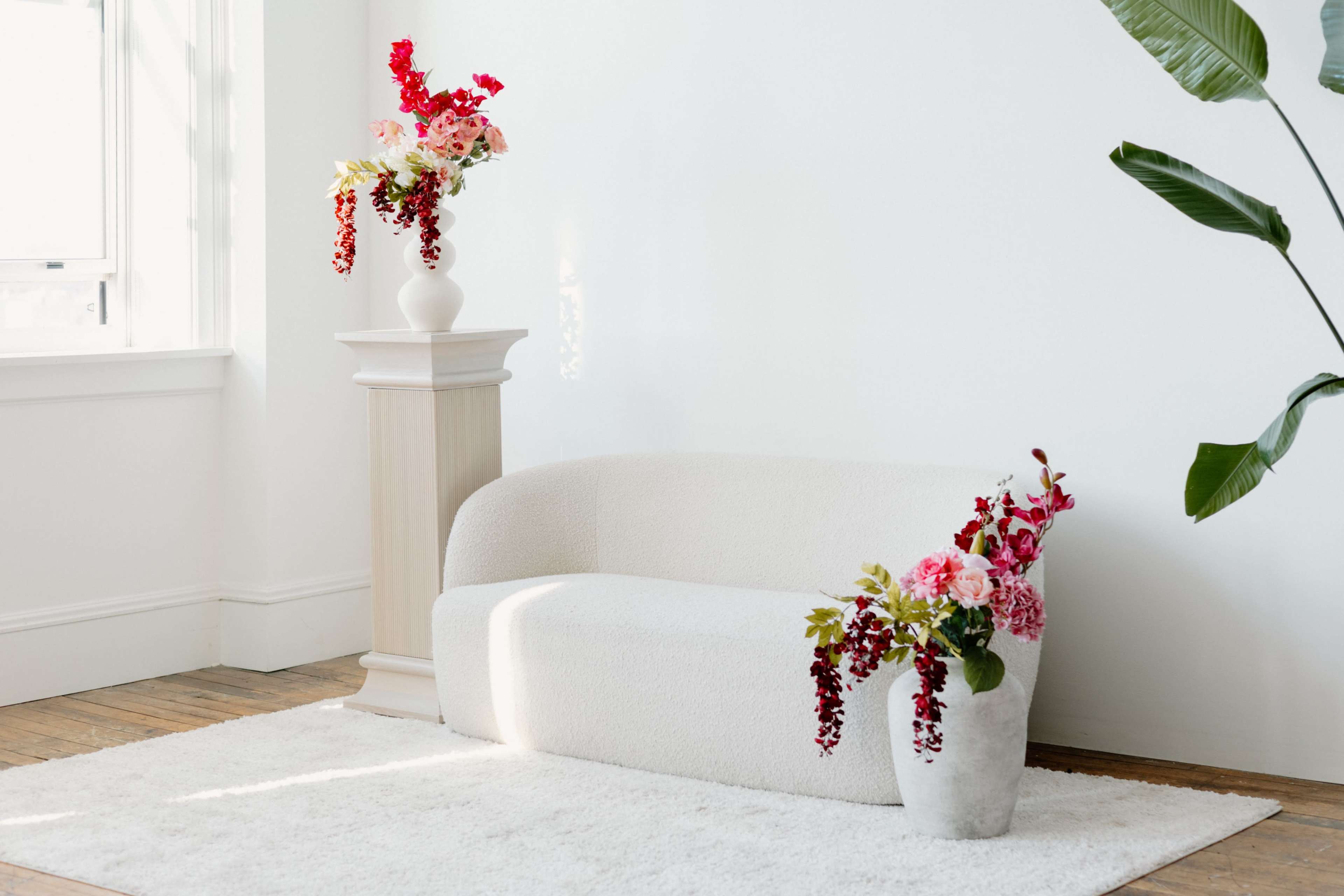 A white sofa is positioned next to a pedestal with a vase of flowers, accompanied by another flower arrangement in a pot on the floor, all set in a bright room with wooden flooring.