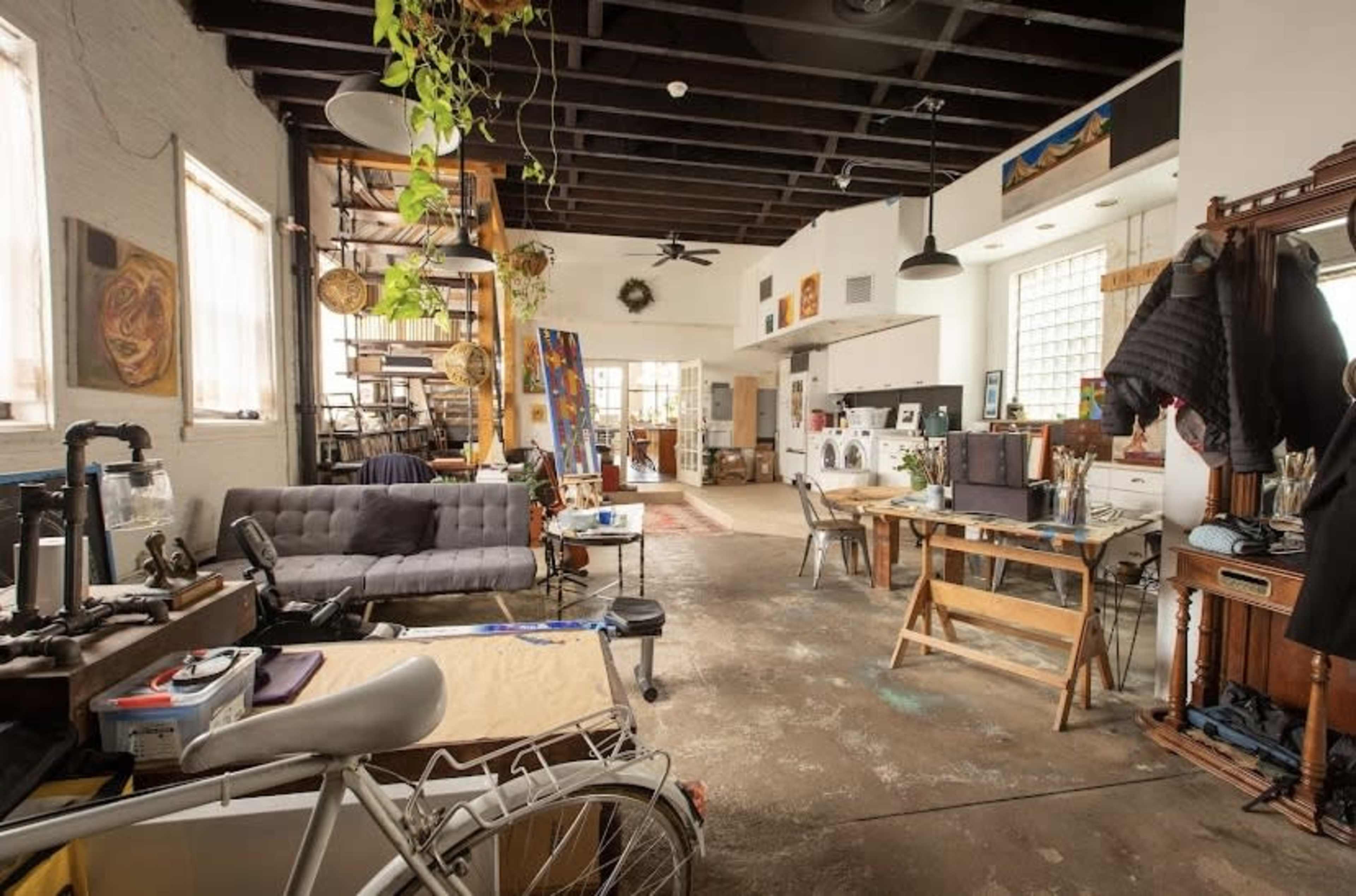 A spacious artist's studio featuring workstations, a sofa, and large windows with various artworks displayed on the walls.