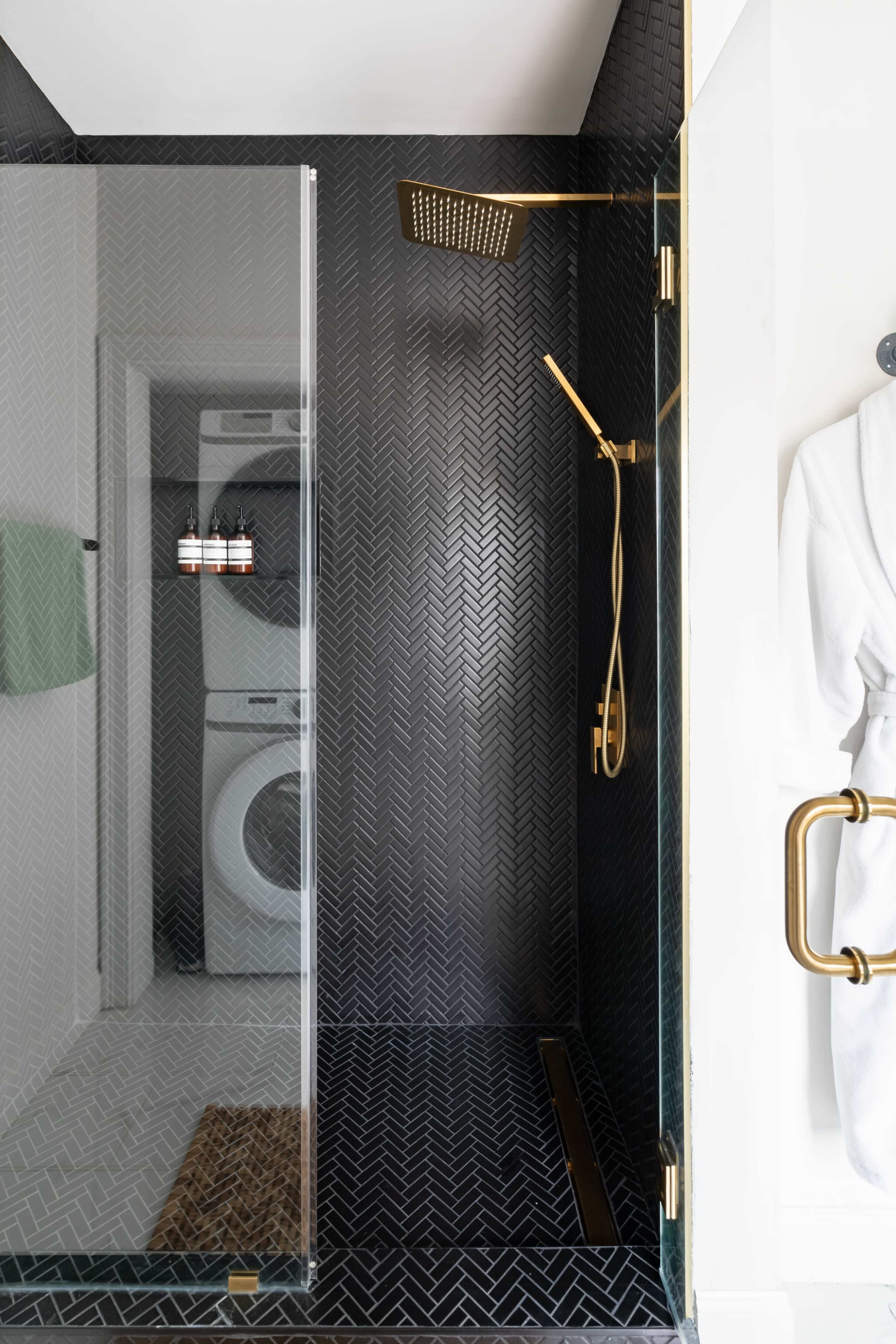 A modern shower with a glass enclosure, a gold showerhead, and a stacked washer and dryer visible in the corner.
