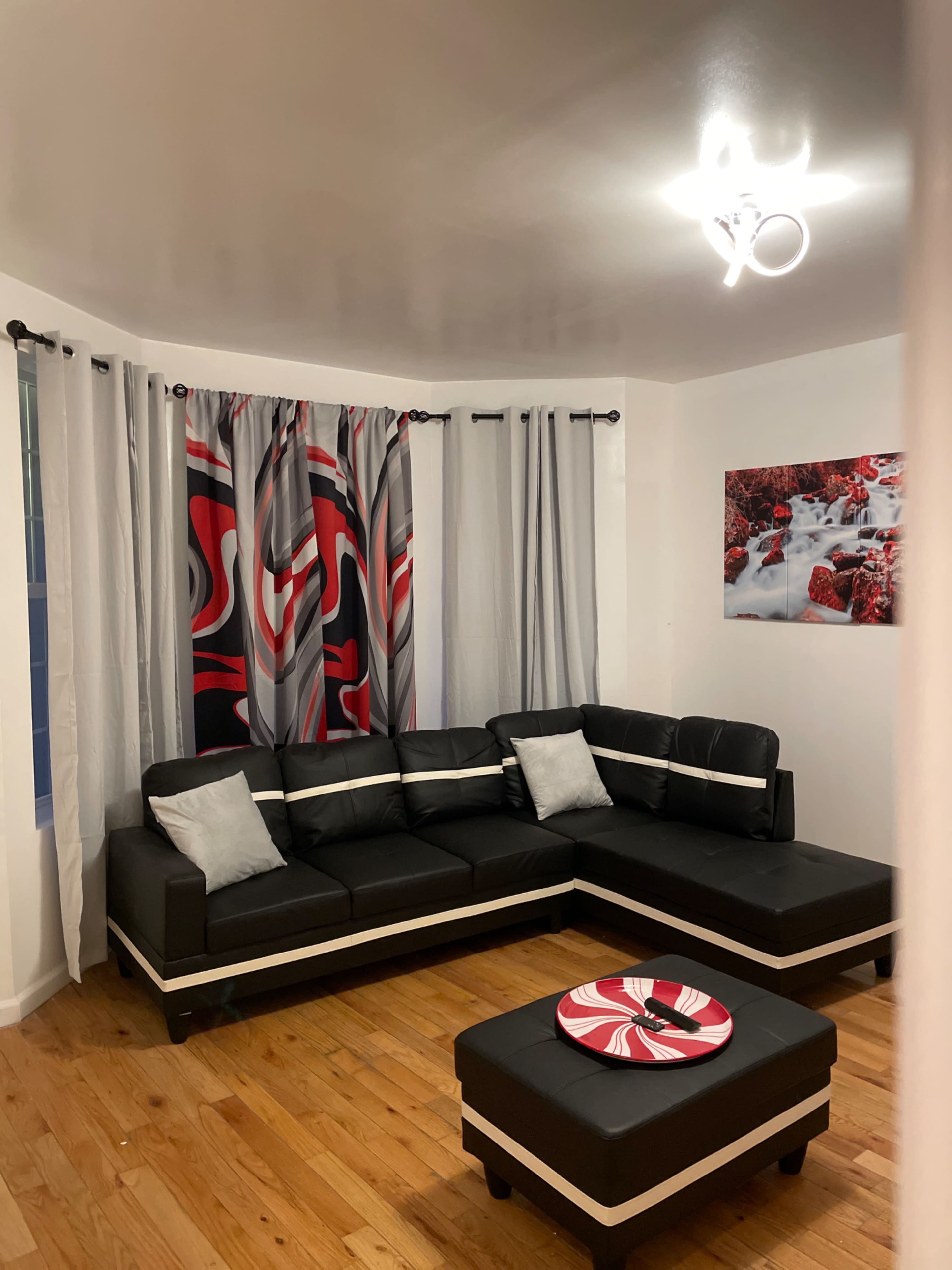 A modern living room with a black sectional sofa, a matching ottoman, curtains with red and gray patterns, and a wall hanging of a water scene.