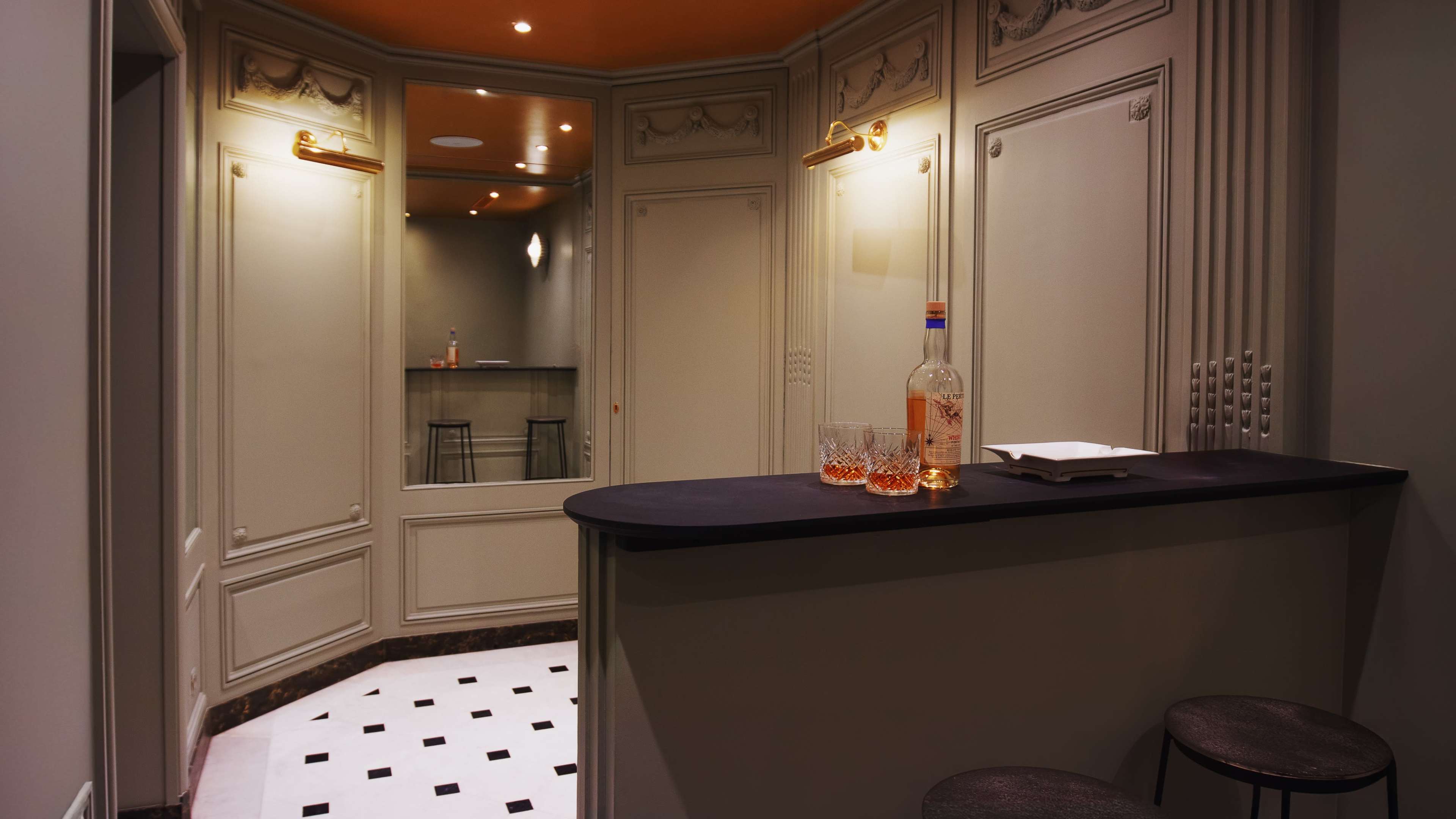 The image depicts a small, elegant bar area with a dark countertop, three stools, and a bottle of liquor alongside glasses on the bar, set against a backdrop of light-colored walls and decorative molding.