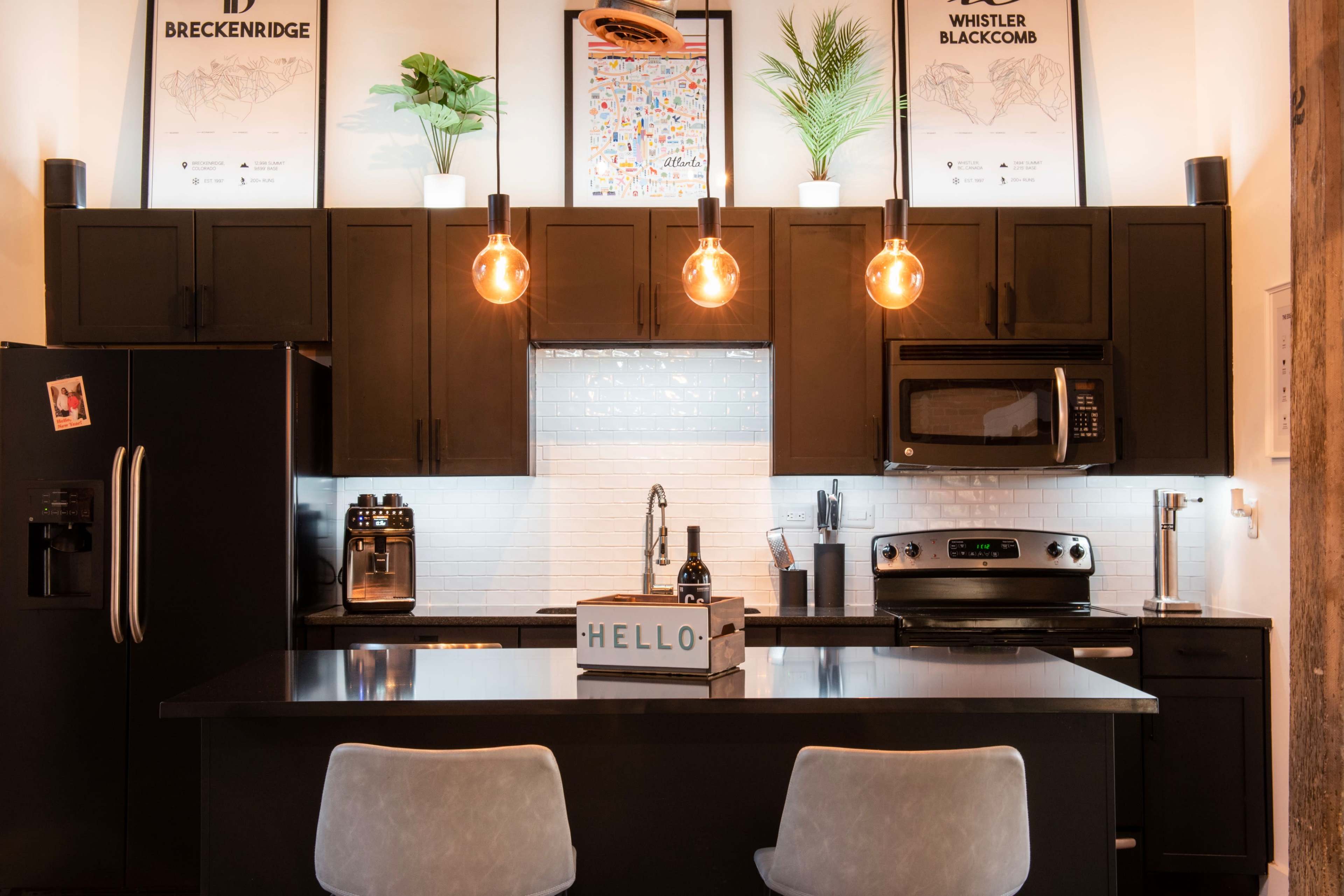 A modern kitchen with black cabinetry, pendant lights, and a central island featuring a wine bottle and a box labeled "HELLO."