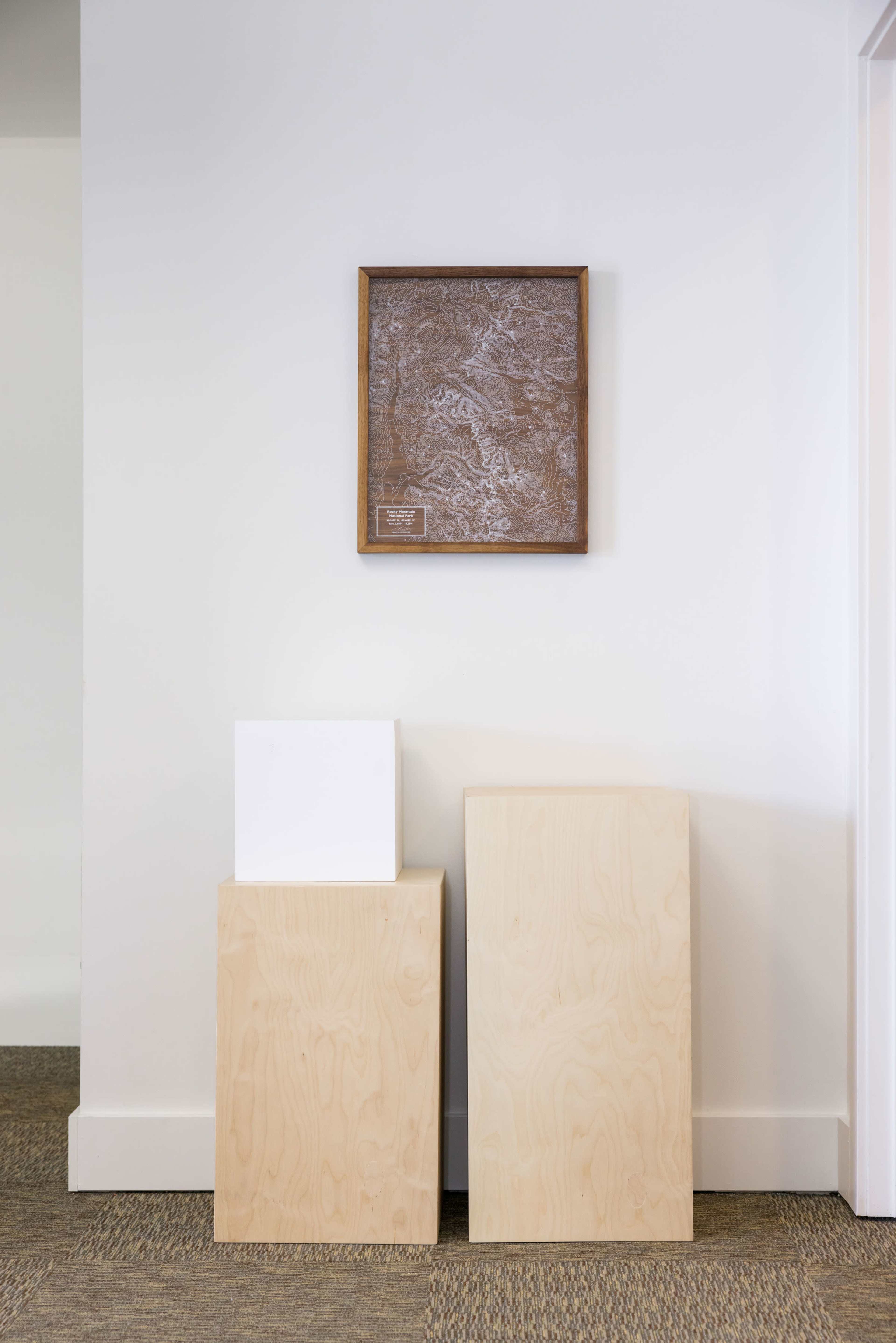 A minimalistic corner of a room with three wooden blocks stacked beside a framed textured artwork on the wall.