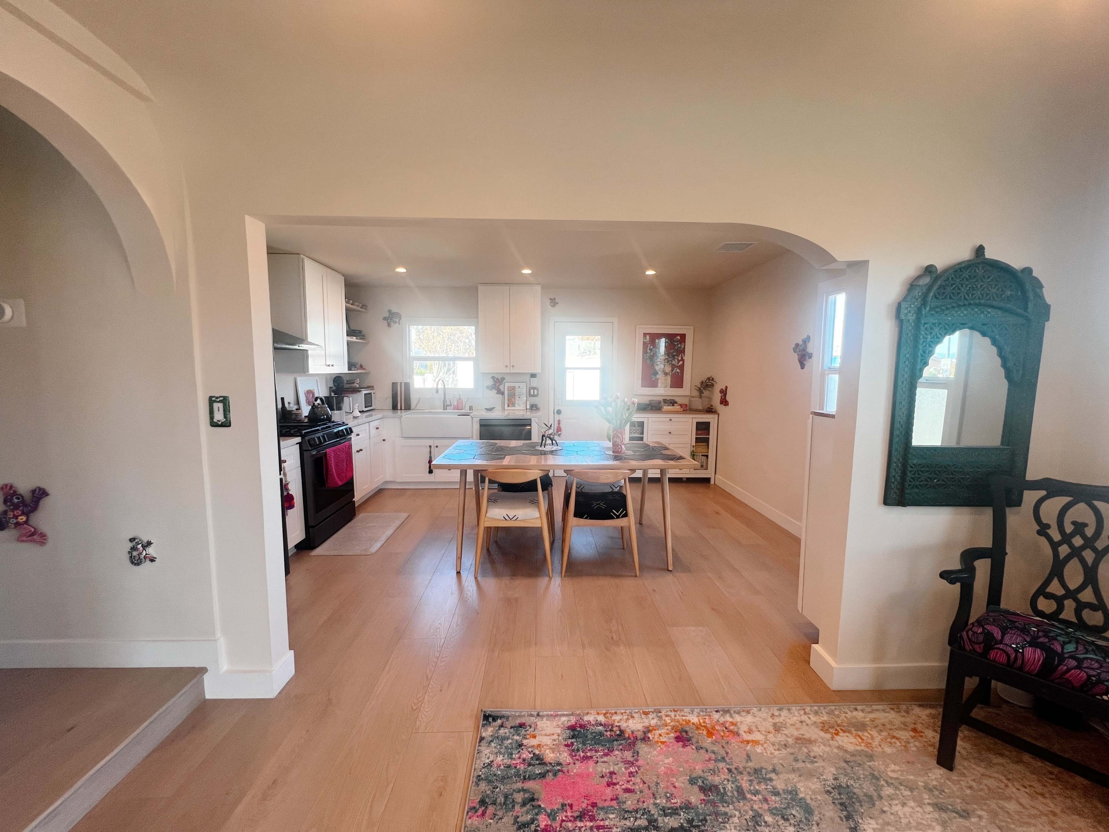 The image shows a modern kitchen and dining area with light-colored walls, wooden flooring, and a table set for dining.