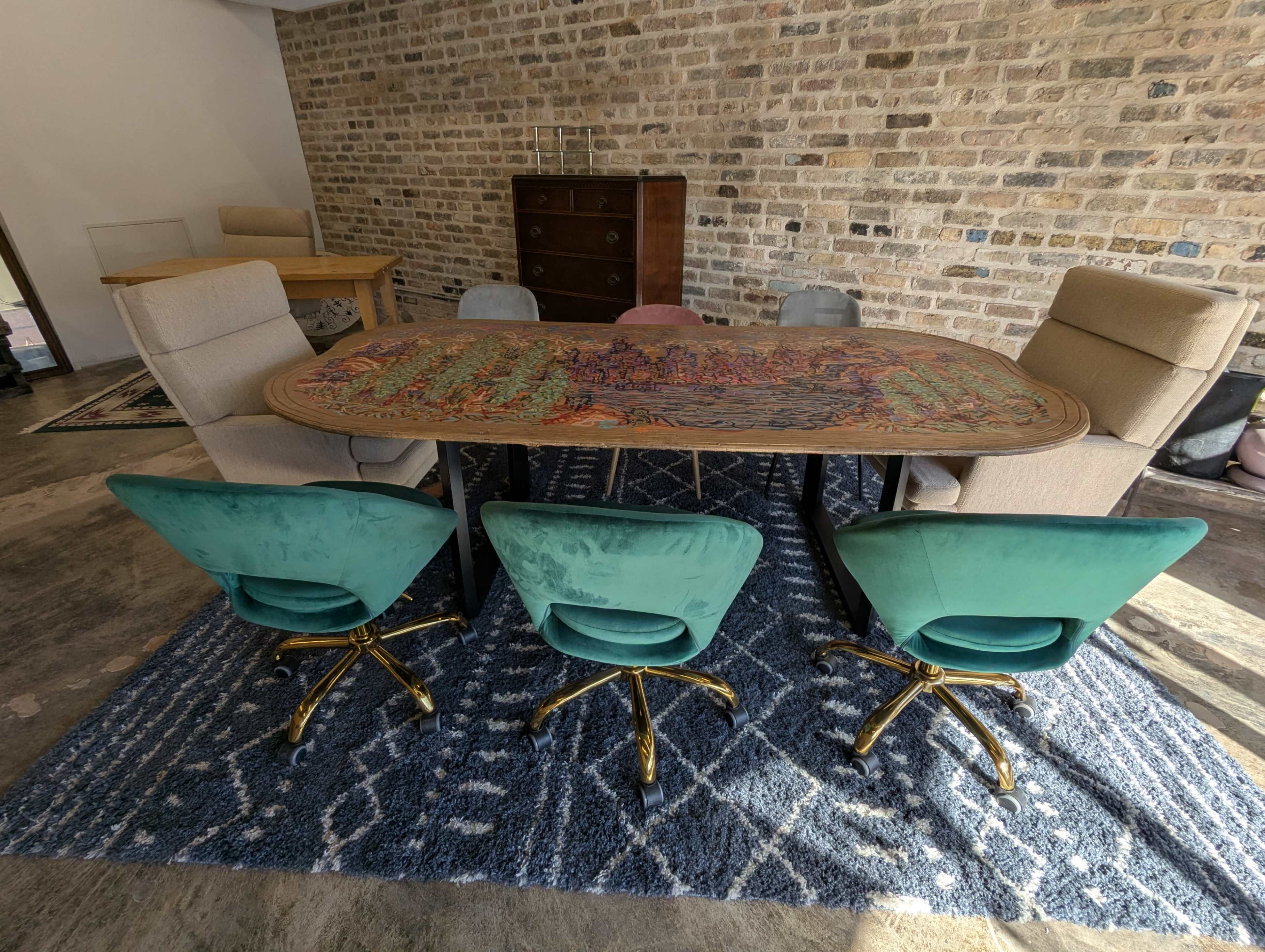 A large, oval table with a colorful design is surrounded by five swivel chairs on a patterned rug in a room with exposed brick walls.