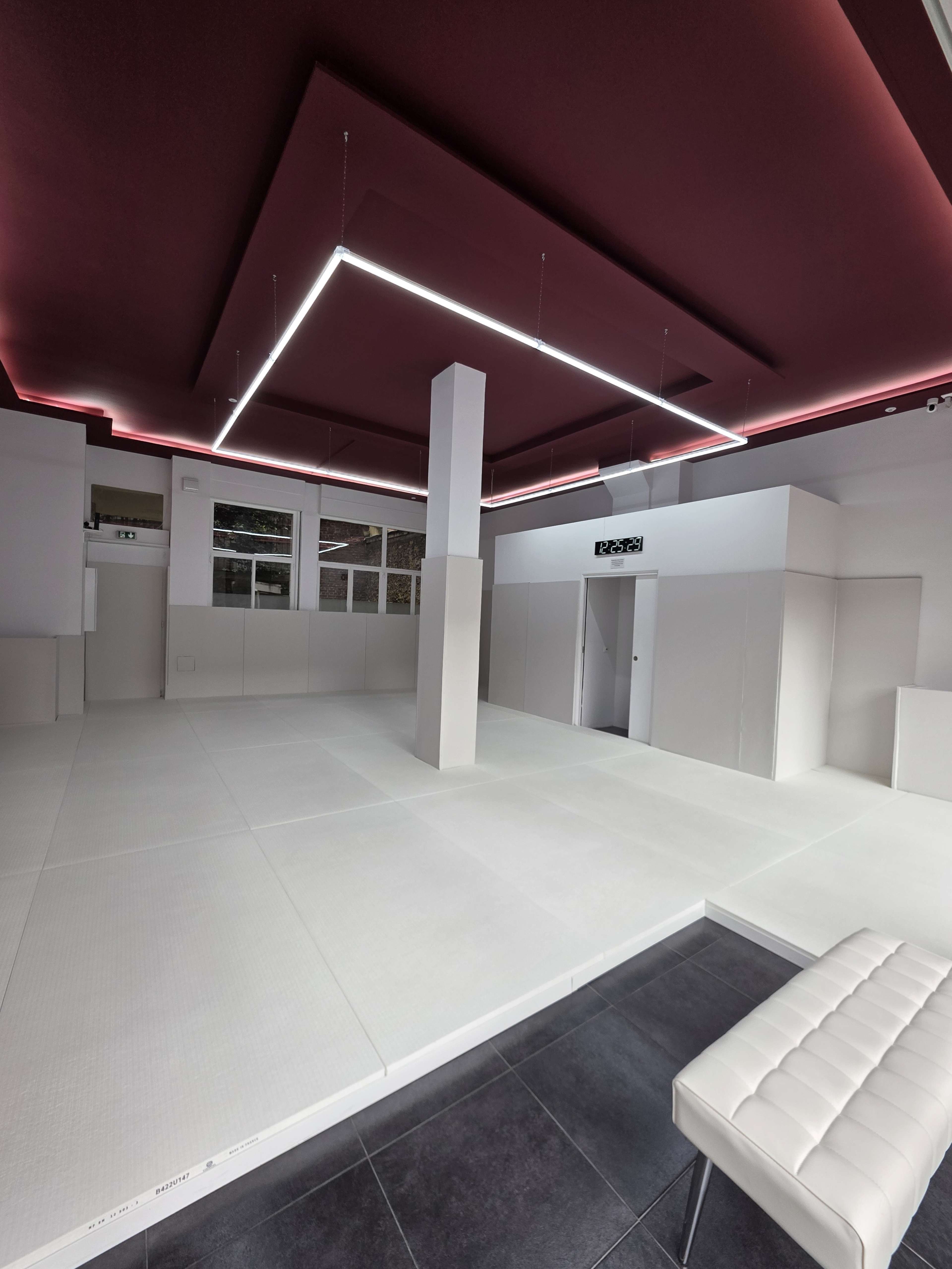 The image shows a spacious, minimalist martial arts dojo with a white tatami mat and a sleek, modern ceiling featuring recessed lighting.