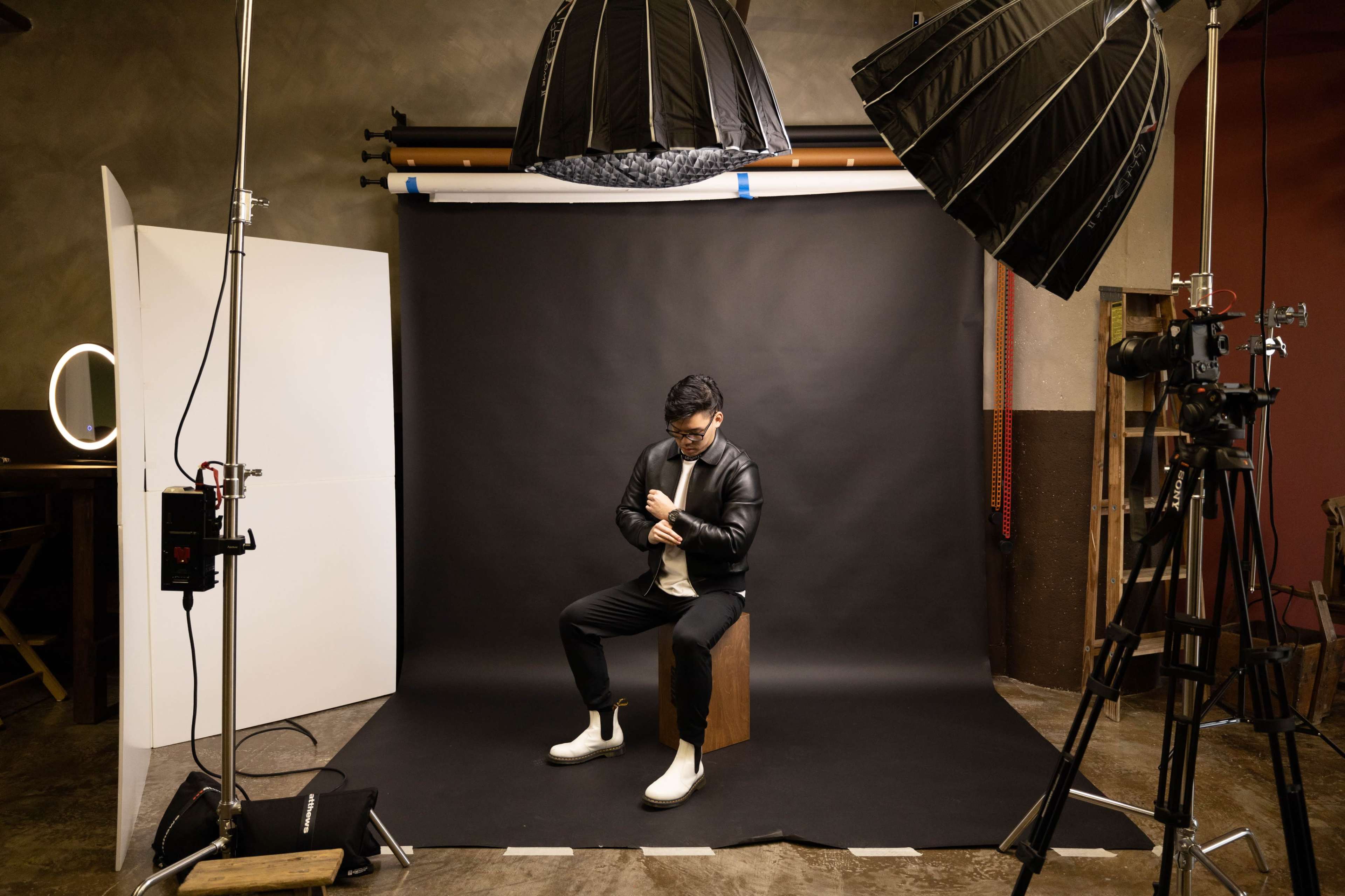 A man wearing a black jacket and white shoes sits on a wooden box in a photography studio, surrounded by lighting equipment and a black backdrop.
